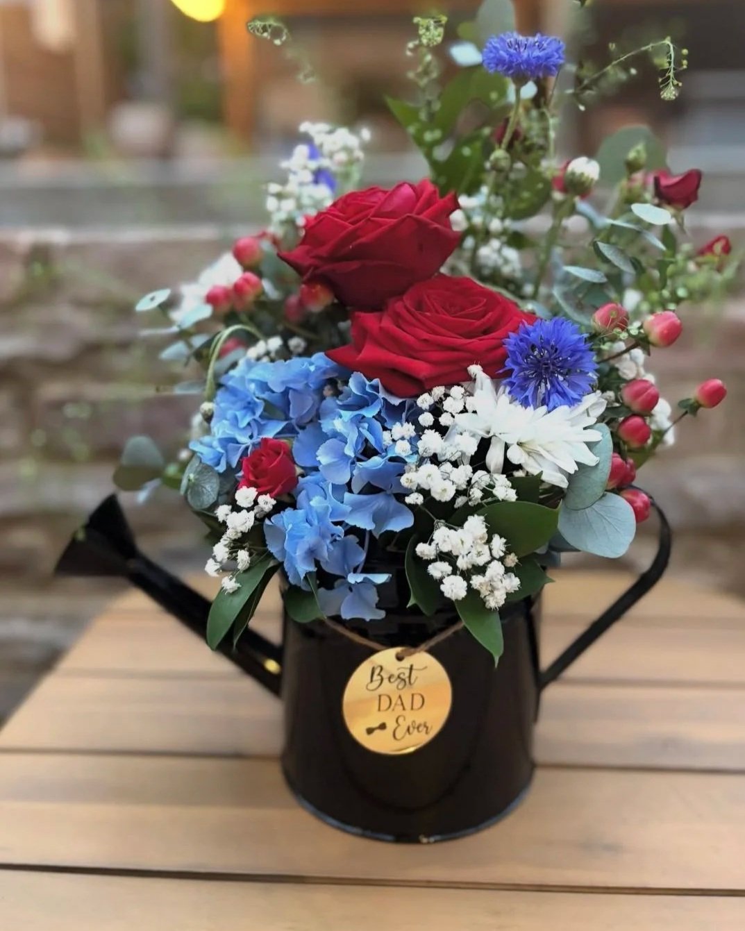 A flower arrangement with red roses, blue hydrangeas, white daisies, baby's breath, and greenery in a black container with a tag that reads 'Best Dad Ever' on a wooden table.