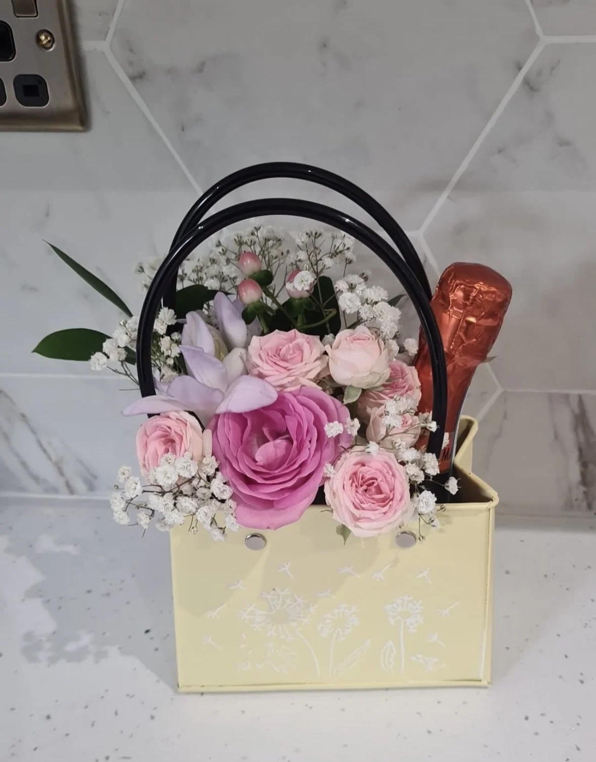 A small bouquet of pink roses, lilacs, white baby's breath, and other flowers in a yellow gift bag with black handles. A wrapped red chocolate bar is placed in the bag.