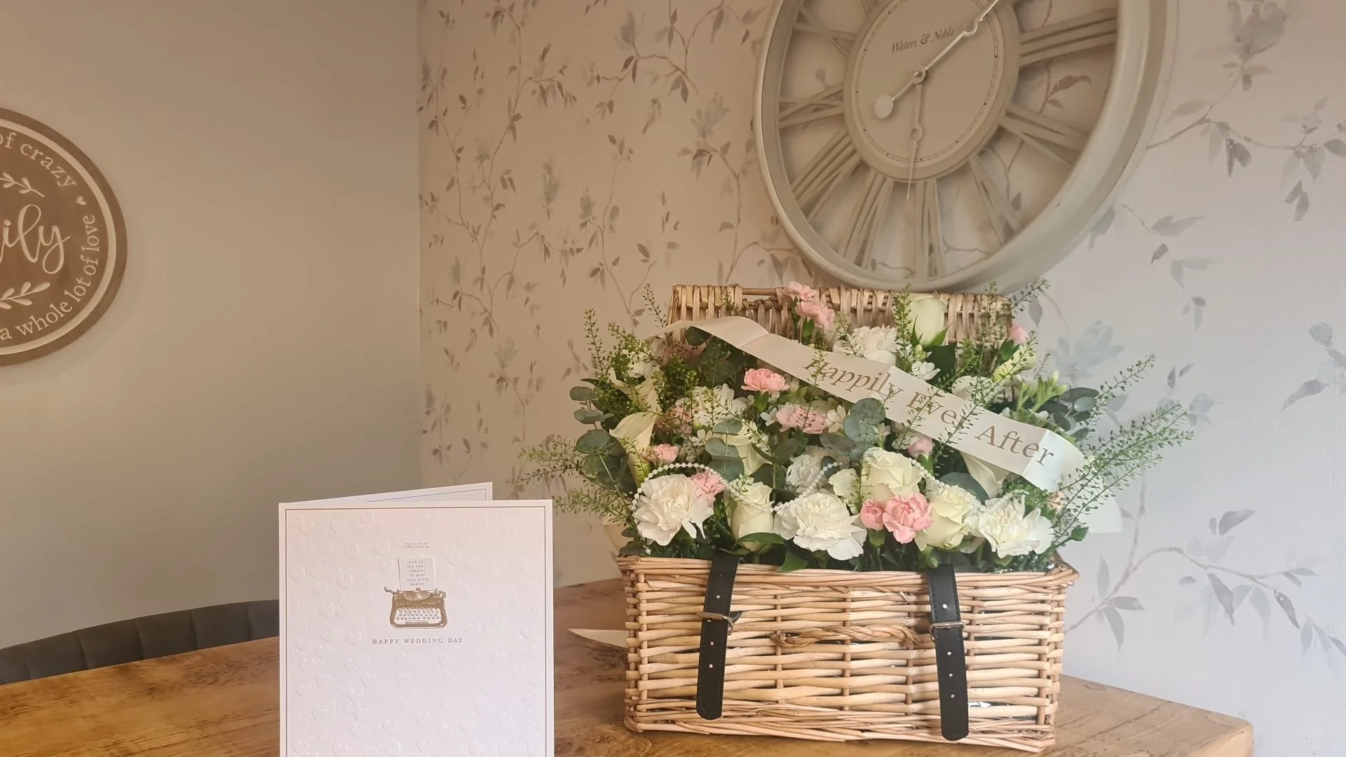 A wicker basket filled with pink and white flowers, placed on a wooden table with a wedding greeting card in front of it. A large wall clock with Roman numerals and a floral wallpaper decorates the background.