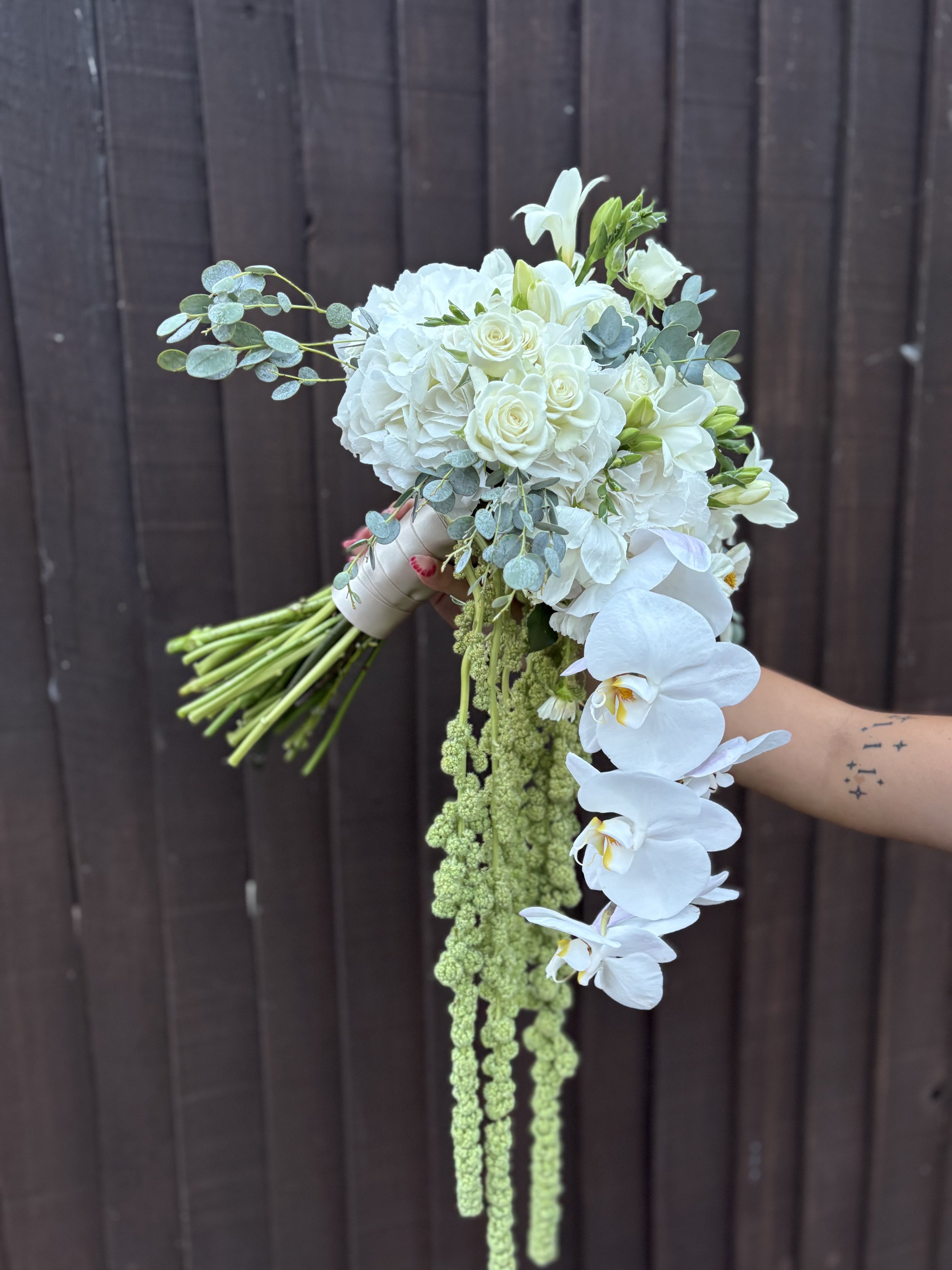 A hand holding a bouquet of white roses, orchids, eucalyptus leaves, and trailing green amaranthus flowers against a dark wooden background.