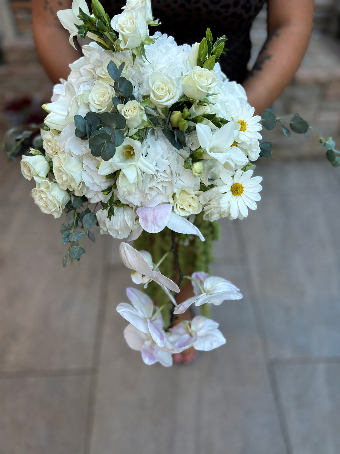A person holding a wedding bouquet of white roses, daisies, calla lilies, and eucalyptus leaves.