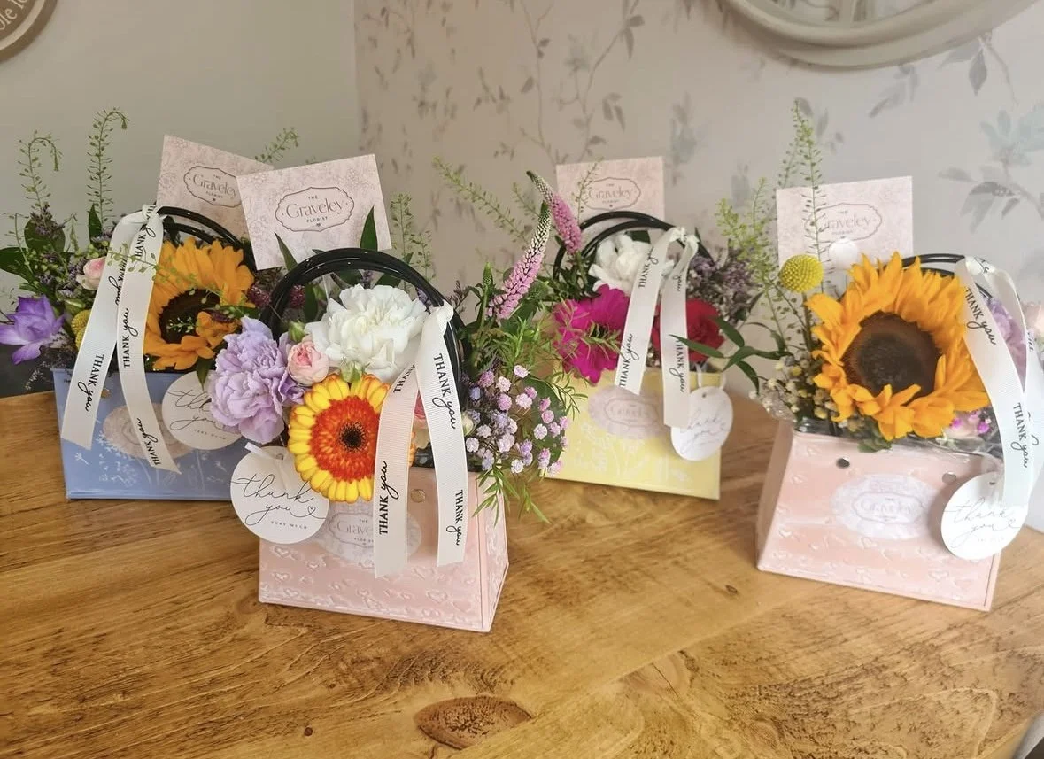 Three decorative floral gift baskets with colorful flowers including sunflowers, roses, and other blooms, each with a ribbon and a "Thank you" tag, placed on a wooden surface.