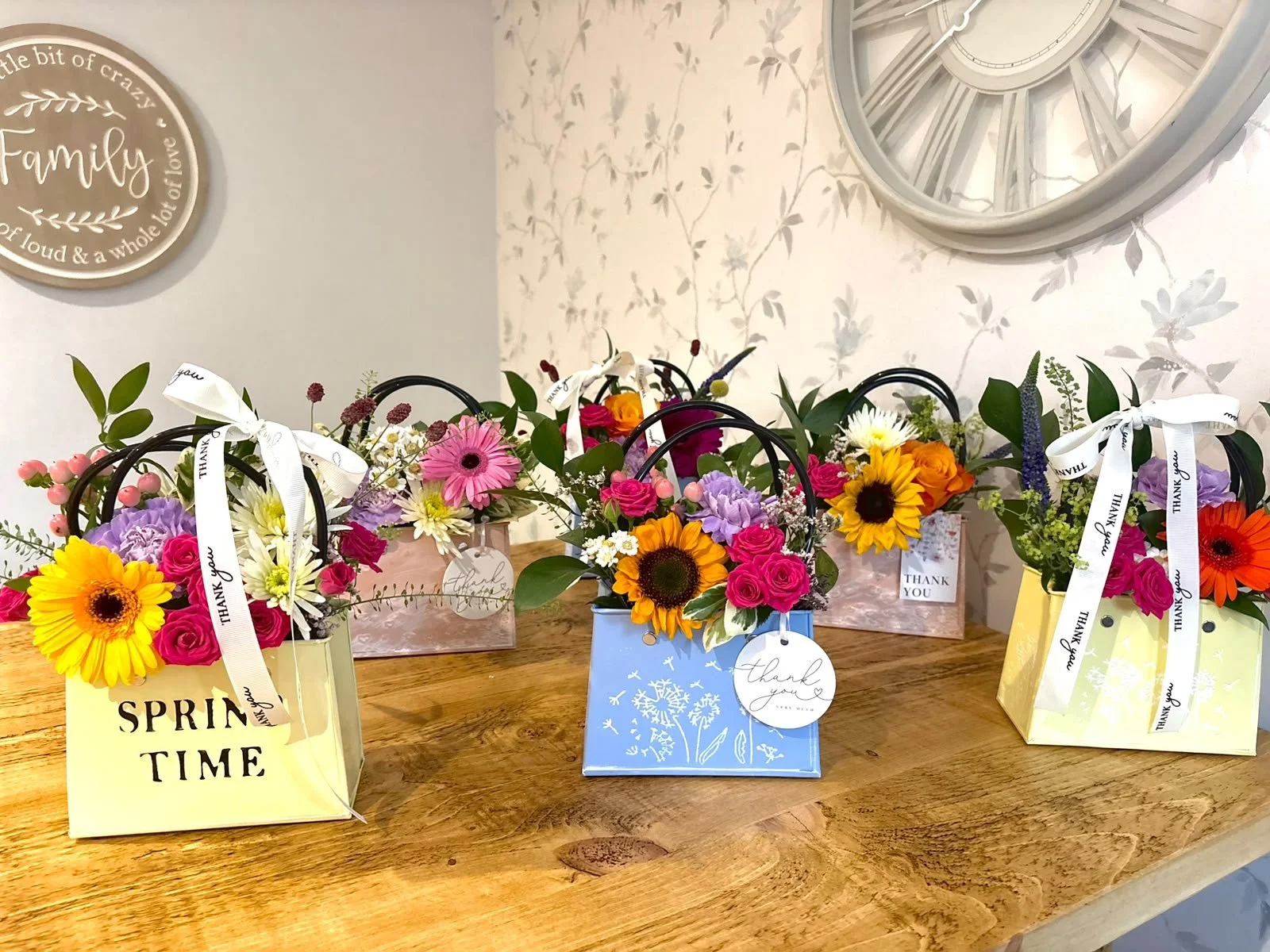 Colorful spring flower arrangements in gift bags and boxes with 'Thank You' tags on a wooden table.