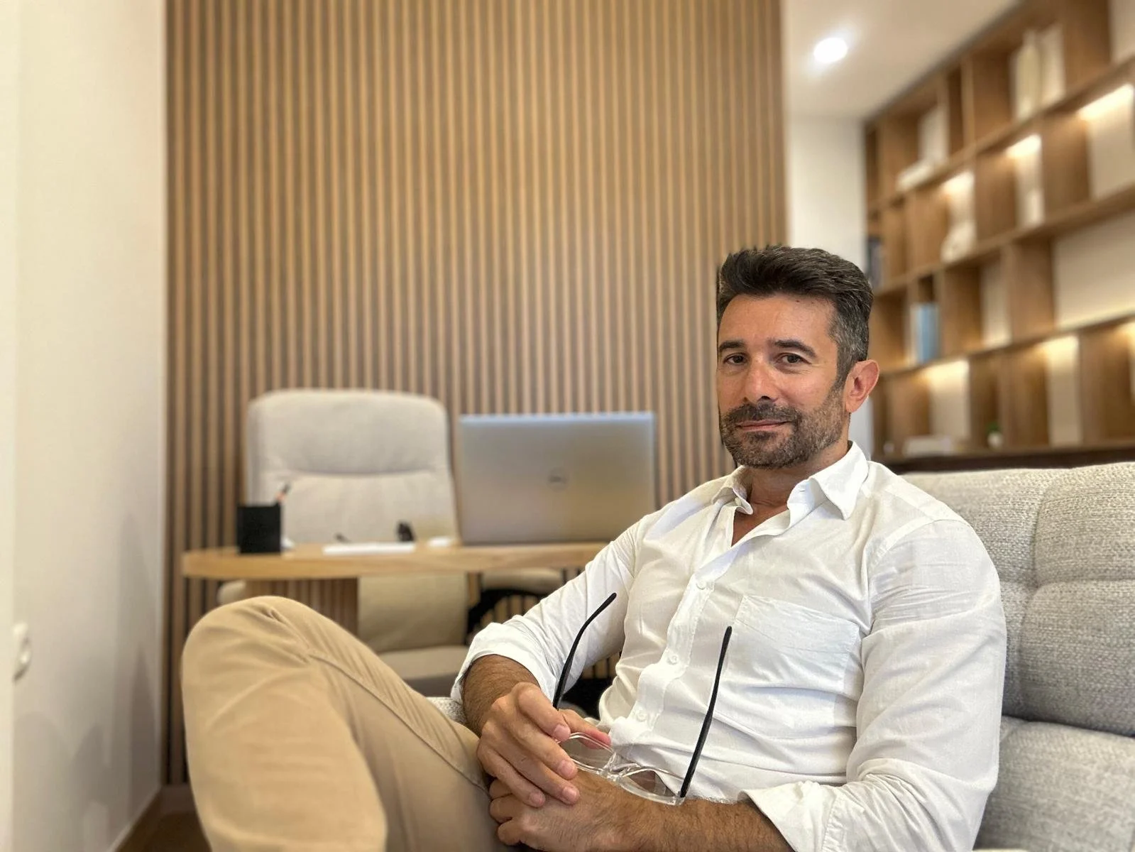 Homem de cabelos escuros e barba, vestindo camisa branca, sentado em um sofá, segurando óculos, em um ambiente com parede de madeira e estantes de livros ao fundo.