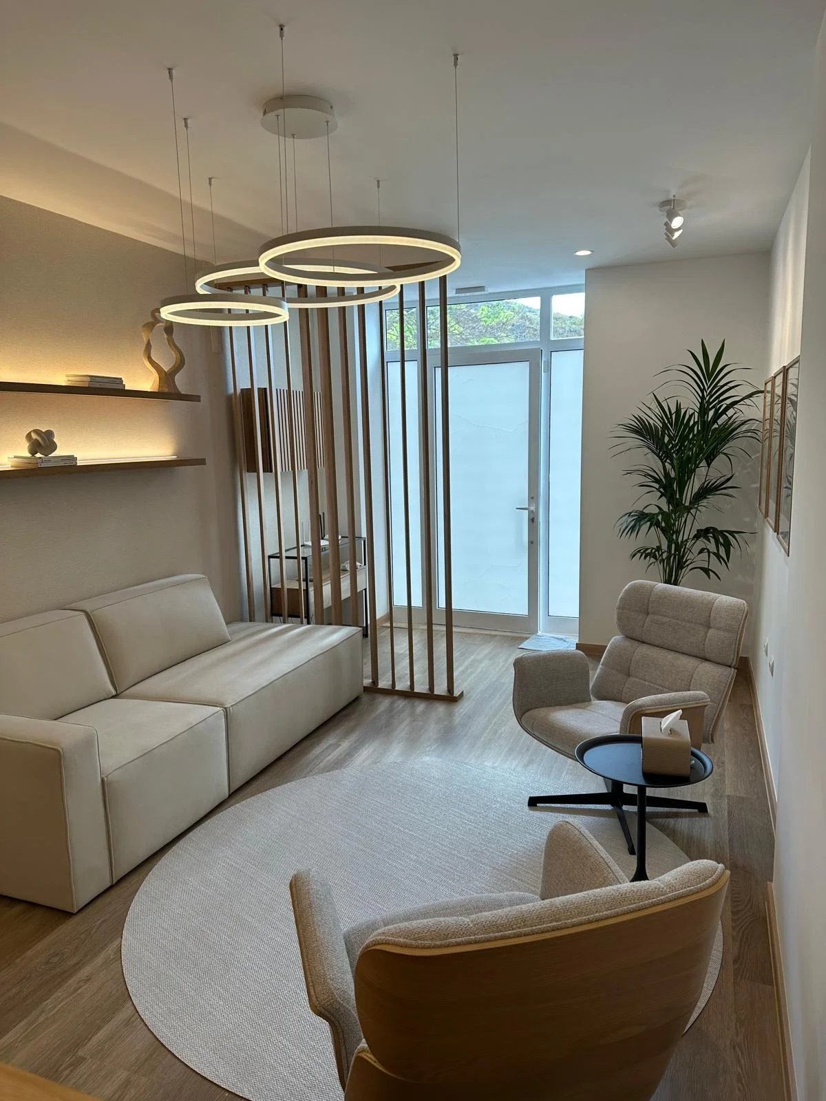 Sala de estar com sofá branco, poltrona cinza, mesa de centro preta, tapete oval branco, prateleira com objetos decorativos, cortina de vidro na porta de saída, luminária moderna no teto, planta grande no canto, paredes brancas, piso de madeira.