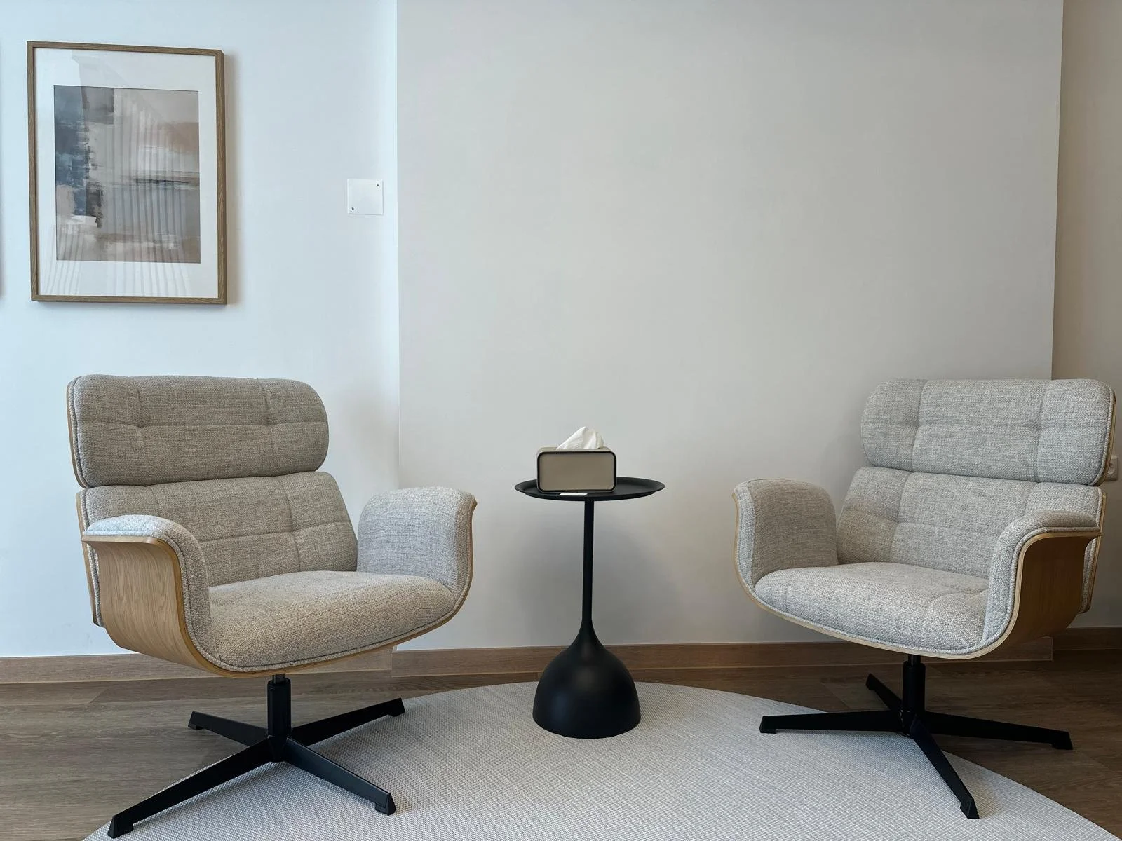 Doisamentos de poltrona em estilo escandinavo com encosto acolchoado, suportes de braço curvos de madeira, um tabuleiro preto com uma caixa de lenços e uma cara de papel, uma parede branca com uma moldura de quadro na parede e um tapete claro sob as cadeiras.