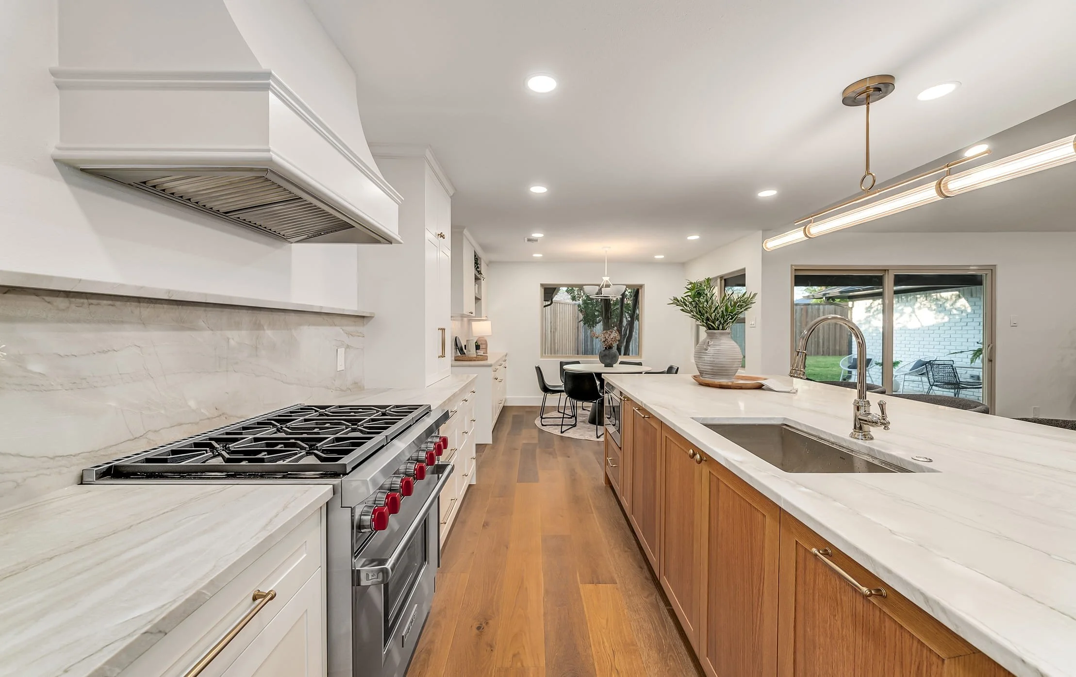 A modern kitchen with white and wooden cabinets, marble countertops, a stainless steel stove, and a large island with a sink. There are large windows and a view of the backyard.