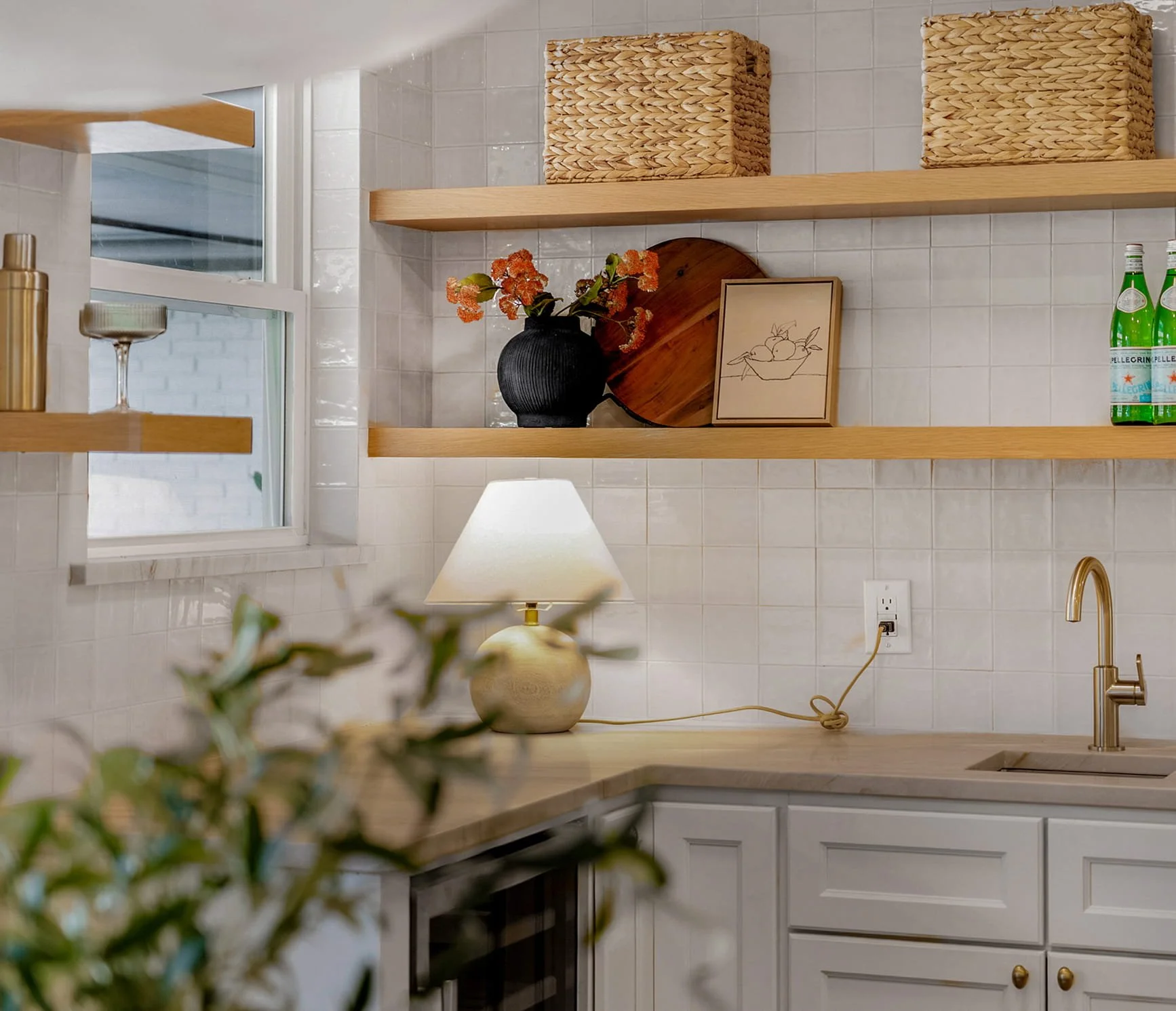 A kitchen with white cabinets, a beige countertop, a gold faucet, and a backsplash of white tiles. There are wooden shelves on the wall holding woven baskets, a black vase with orange flowers, a framed drawing, and green bottles. A white table lamp and a power outlet are also visible.