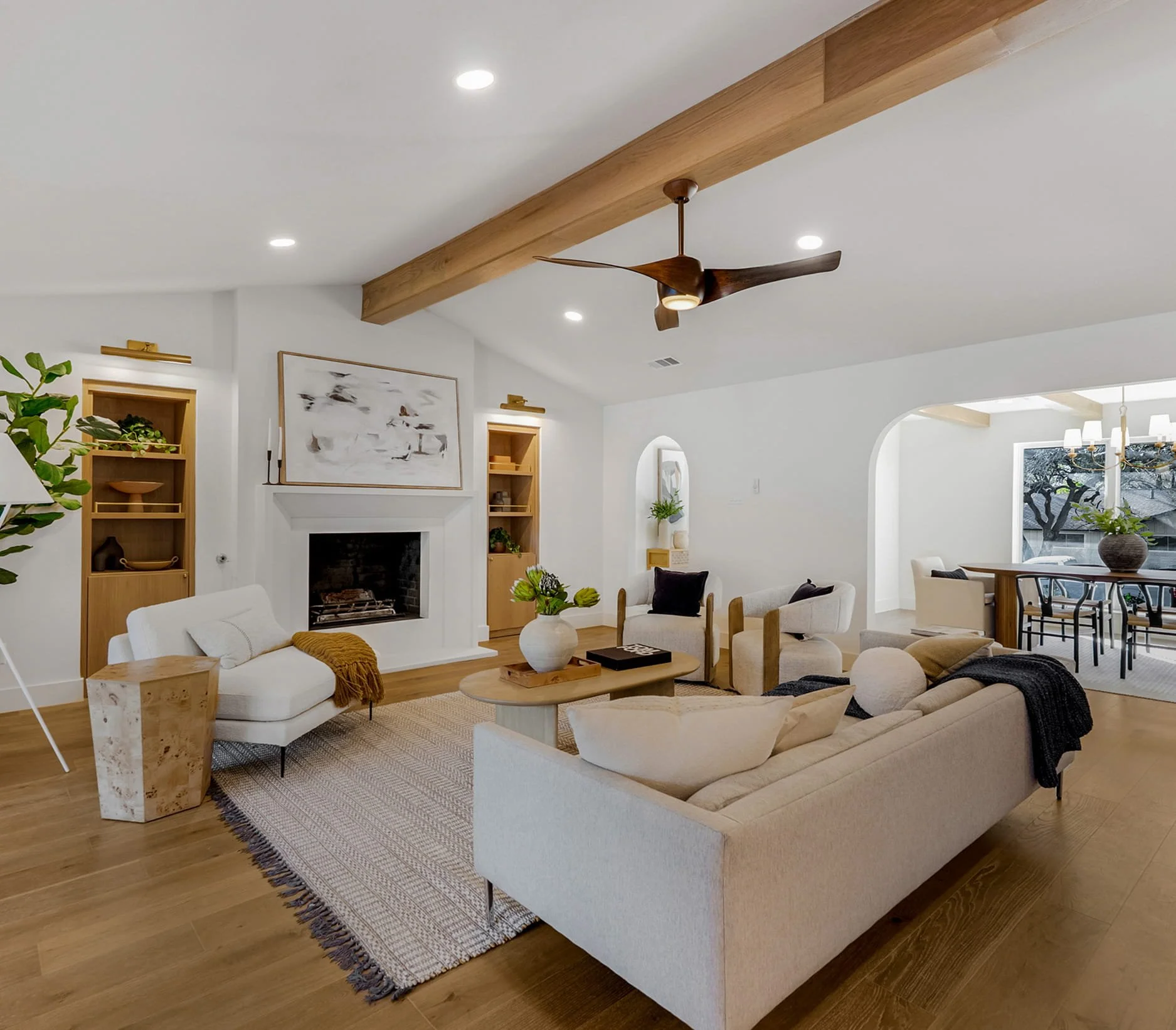 A spacious, modern living room with white walls, hardwood floors, and a white fireplace. There are multiple beige and cream-colored sofas and chairs, a wooden coffee table, and decorative plants. A wooden beam runs across the ceiling with a mounted ceiling fan. Large windows let in natural light, and the room features minimalist artwork and built-in wooden shelves.