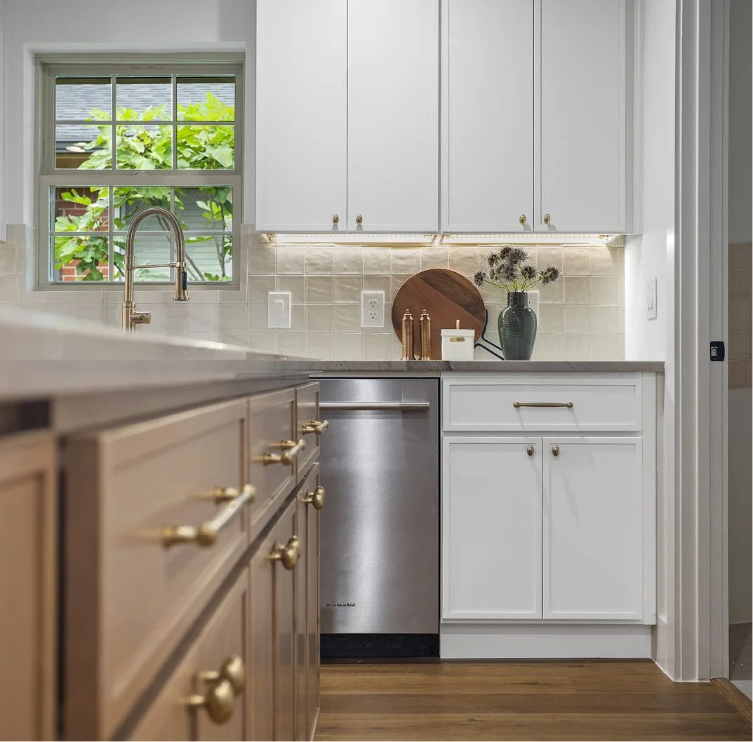 A well-lit kitchen featuring a window with green foliage outside, white cabinetry with gold hardware, beige tile backsplash, a stainless steel dishwasher, and decorative items including a vase with flowers, cutting boards, and gold containers.