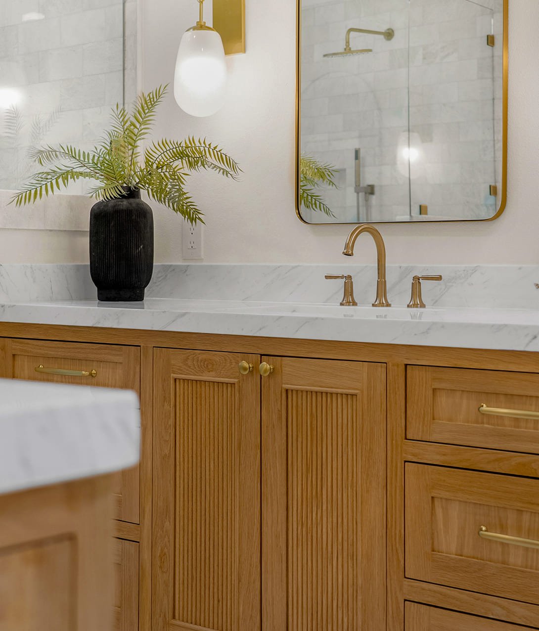 Bathroom vanity with a wooden cabinet, a white marble countertop, a gold faucet, a mirror, a black vase with green fern leaves, and a wall-mounted white light fixture.