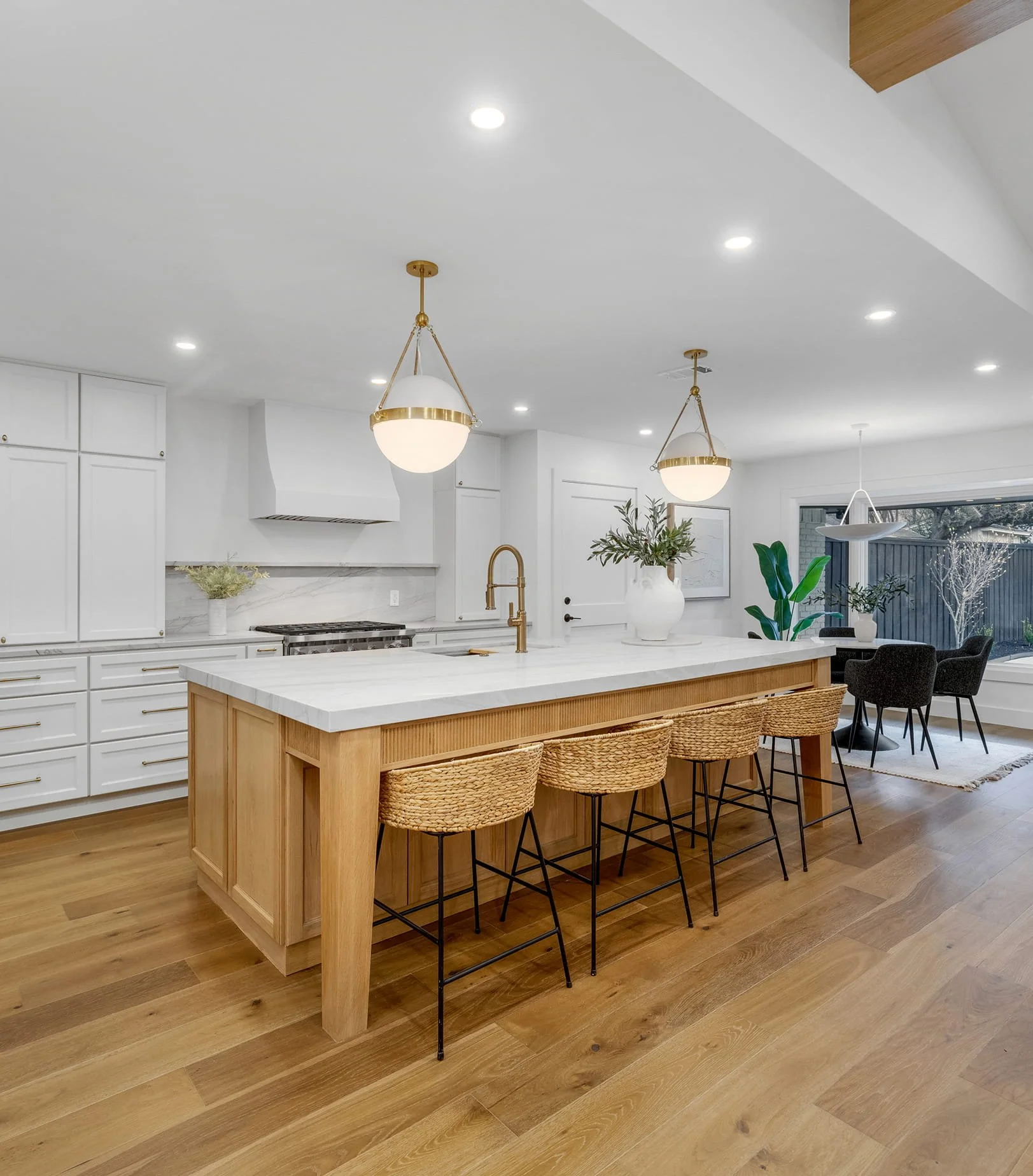 Modern kitchen with white cabinets, a wooden island with four woven rattan chairs, two pendant lights with white shades and gold accents, a marble backsplash, potted plants, and large windows with a view of a deck and trees outside.
