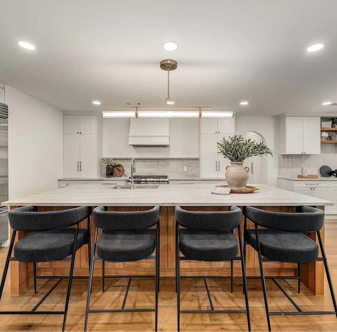 Modern kitchen with white cabinetry, marble backsplash, and a large island with a white countertop. Four black barstools with padded seats line the front of the island. A large potted plant on a tray is on the island. Recessed lighting and a ceiling fixture light the space, with wooden flooring.