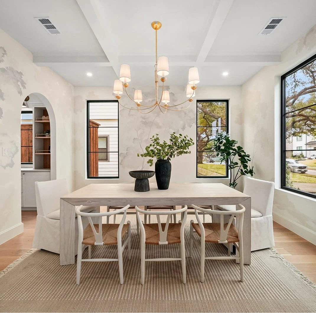 A bright dining room with a rectangular white table, surrounded by six chairs and two armchairs. A black vase with greenery and a black bowl are on the table. Three large windows let in natural light, with a view of the outside yard. A modern chandelier with multiple small lamps hangs above the table. The room has light-colored walls, a white ceiling with cove moldings, and a woven area rug under the table.