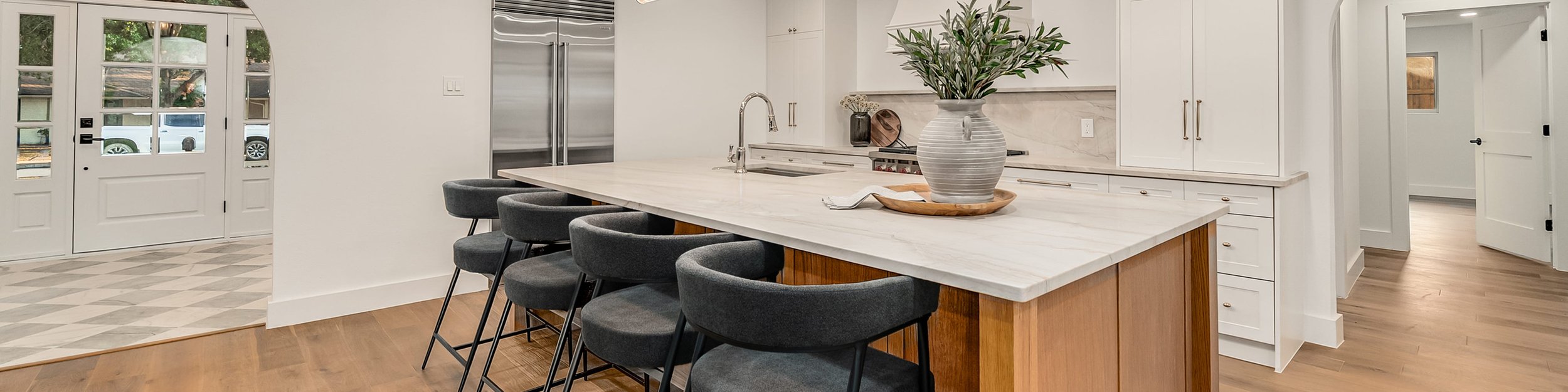 Modern kitchen with white cabinets, marble countertops, stainless steel refrigerator, and a wooden kitchen island with five dark gray bar stools. There is a large vase with greenery on the island.