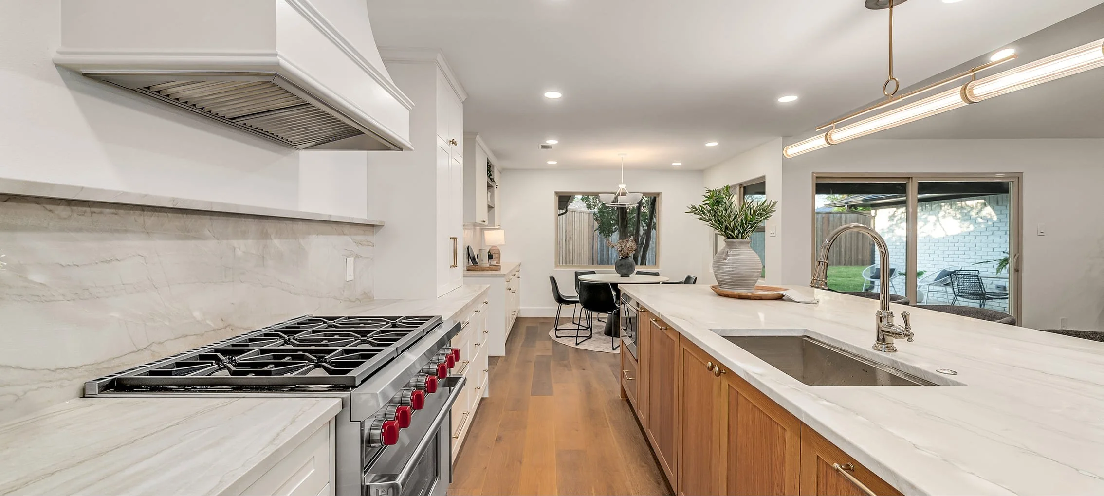 Modern kitchen with white and wood cabinets, marble countertops, a stainless steel stove with red knobs, and a large island with a sink and a potted plant. In the background, there is a dining area with a round table, chairs, and a large window and sliding glass doors showing a backyard with patio furniture.