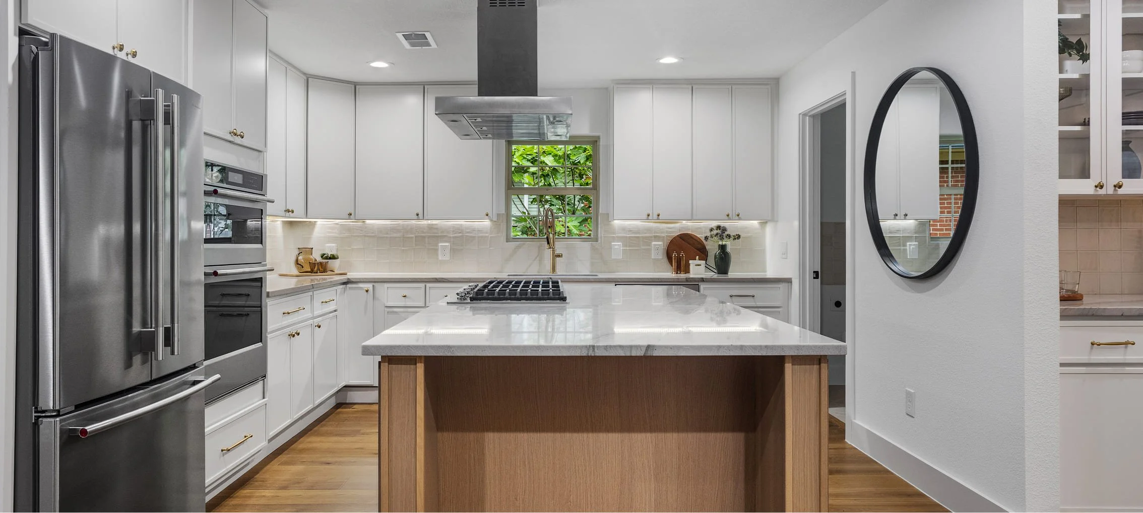 Modern kitchen with white cabinets, stainless steel appliances, a kitchen island with a light-colored countertop, a window showing green foliage, and a round mirror on the wall.