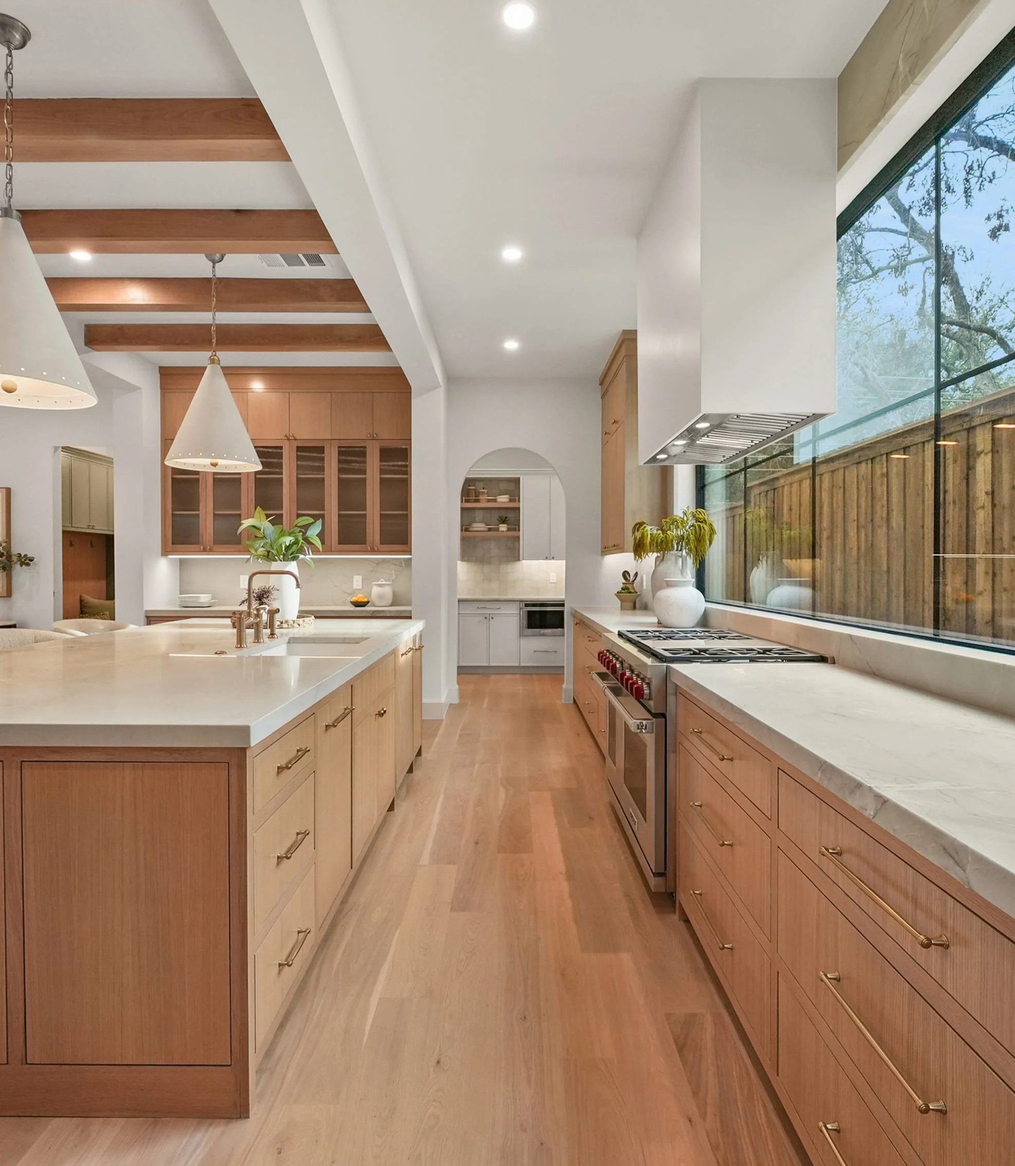 Modern kitchen with wooden cabinets and drawers, white marble countertops, hanging pendant lights, large window with trees outside, and hardwood floors.