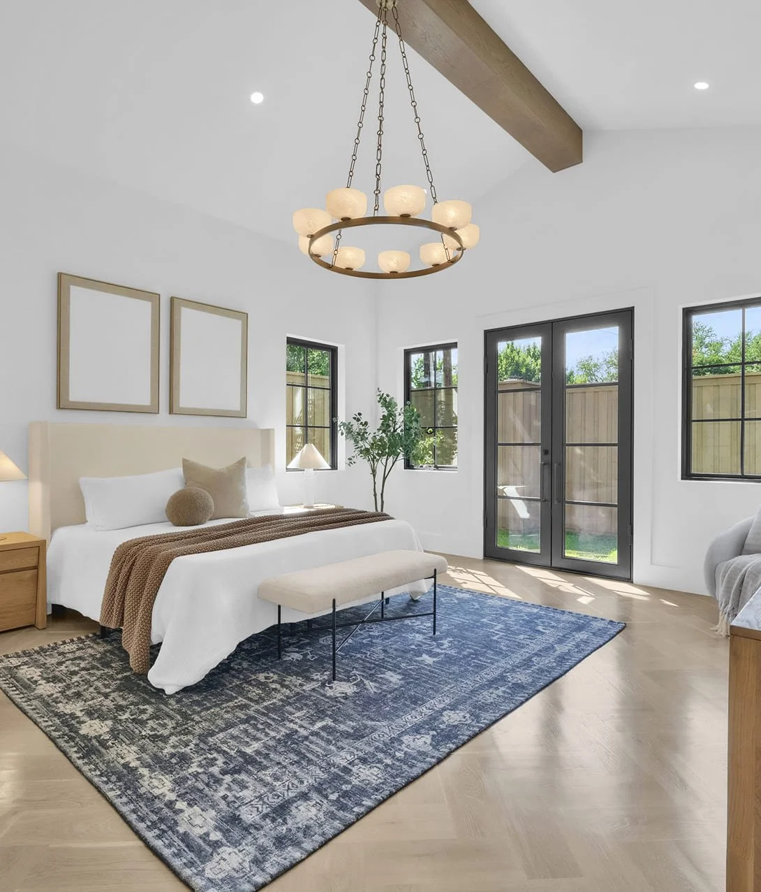 Bright bedroom with a large bed, beige headboard, and decorative pillows. Nightstand with lamp on each side. Blue patterned area rug, benches, minimalist art, and large windows with black frames. Glass door leading outside, ceiling light fixture.