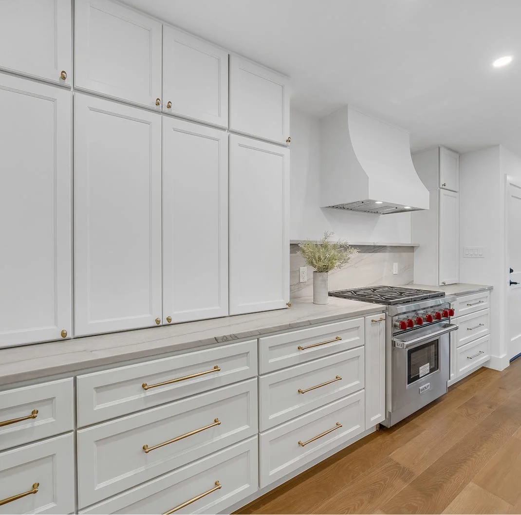 Modern kitchen with white cabinets, stainless steel oven, and gold handles, featuring a wooden floor and a white range hood.