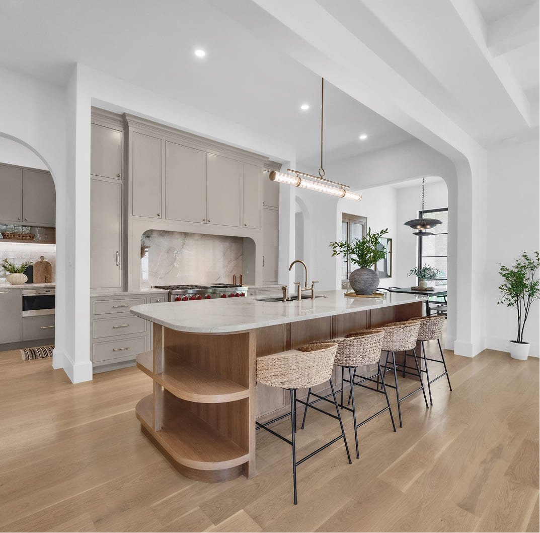 Modern kitchen with an island, beige cabinets, marble backsplash, and bar stools with woven seats, wood flooring, and potted plants.