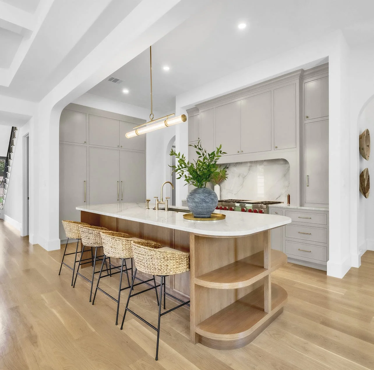 Modern kitchen with white cabinets, marble backsplash, light wooden island, four woven bar stools, gold fixtures, and a potted plant in a gray vase.