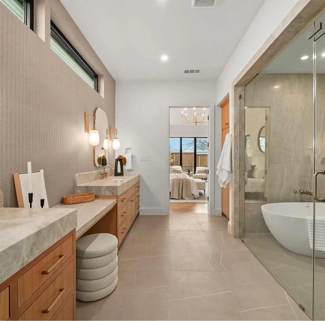 A spacious bathroom with a beige tiled floor and a double vanity with beige counters and wooden cabinets. There are sconces above the vanity mirror, and stacked round ottomans are beside it. The glass shower enclosure has a modern bathtub inside, and a towel hangs on a hook on the wall. Beyond the bathroom, a bedroom with a bed, window, and chandelier is visible.