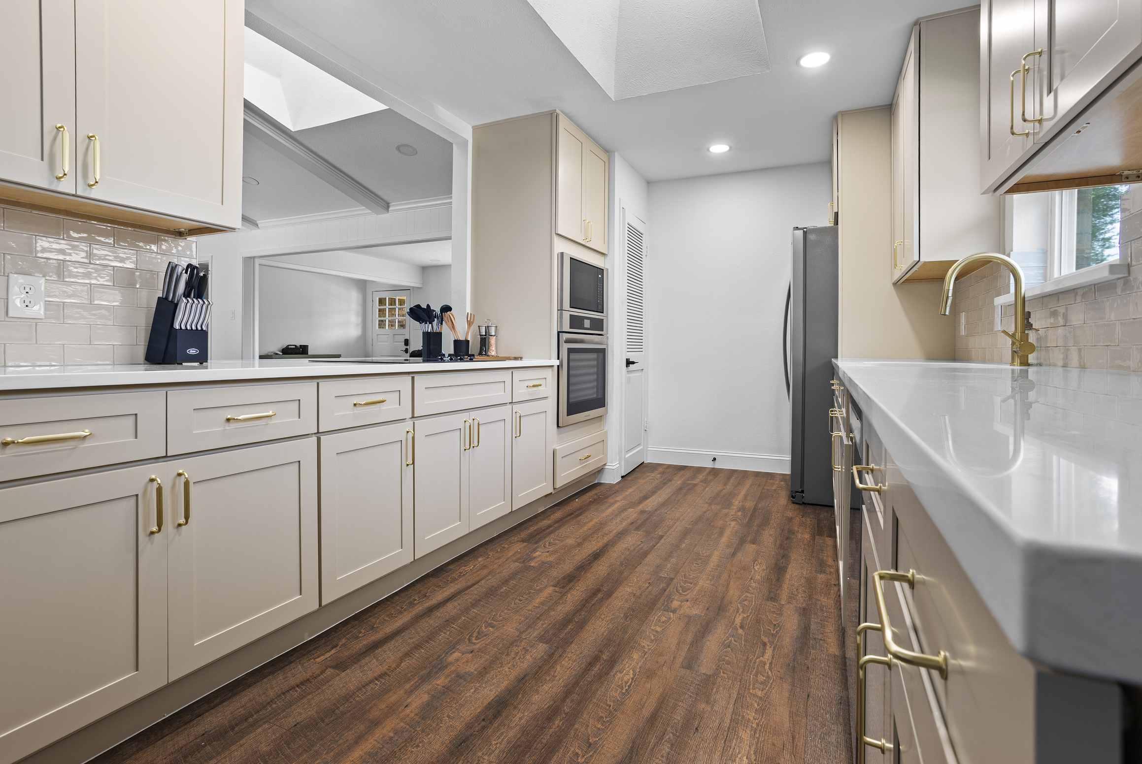 A modern kitchen with white cabinets, beige subway tile backsplash, and wooden flooring. There are built-in appliances, a window above the sink with gold fixtures, and kitchen utensils on the counter.