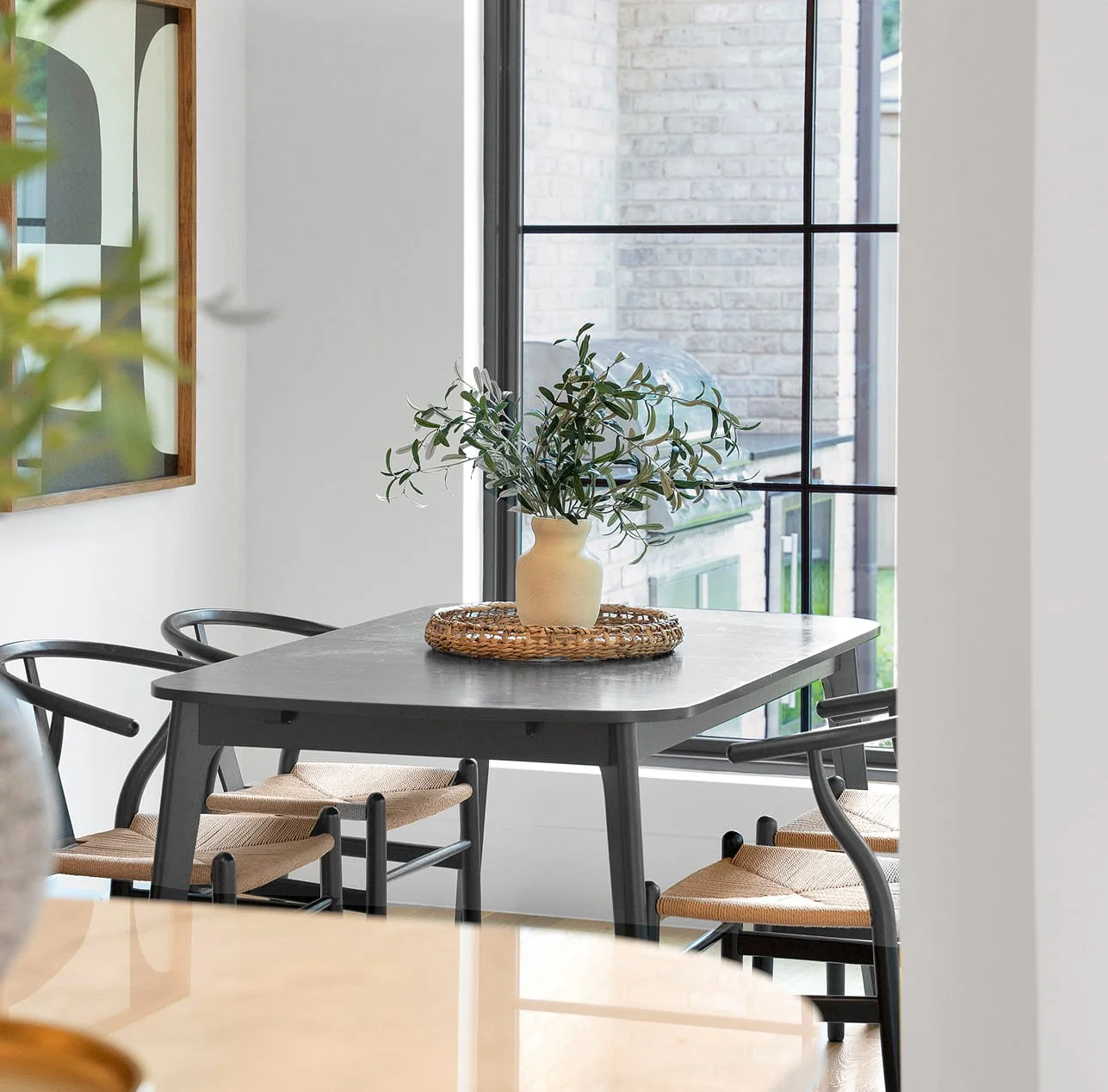 Dining area with a table, a potted plant in a cream vase on a woven tray, and chairs, next to a large window showing a brick exterior.