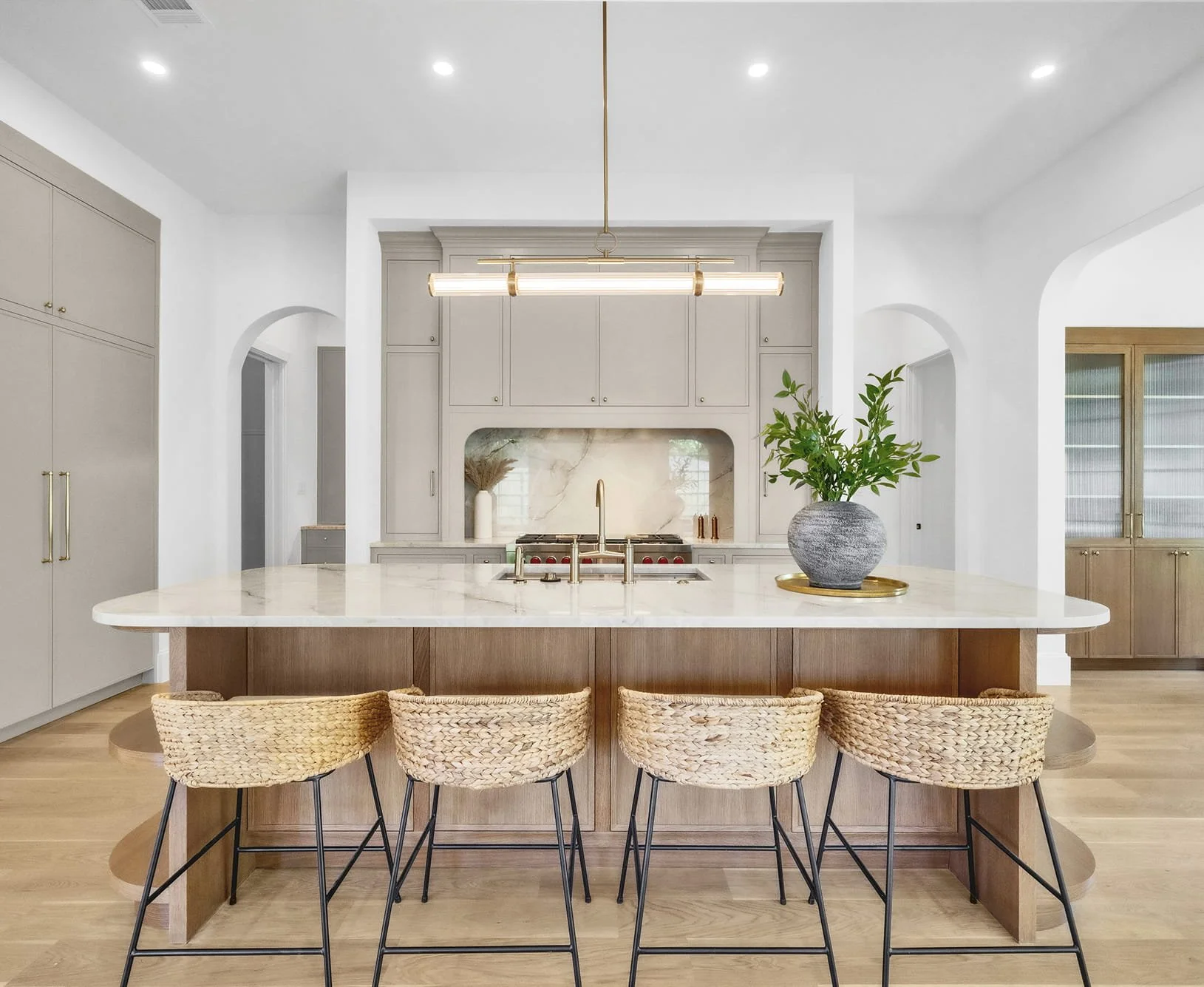 Modern kitchen with island, wicker chairs, beige cabinetry, white walls, and a potted plant on marble countertop.