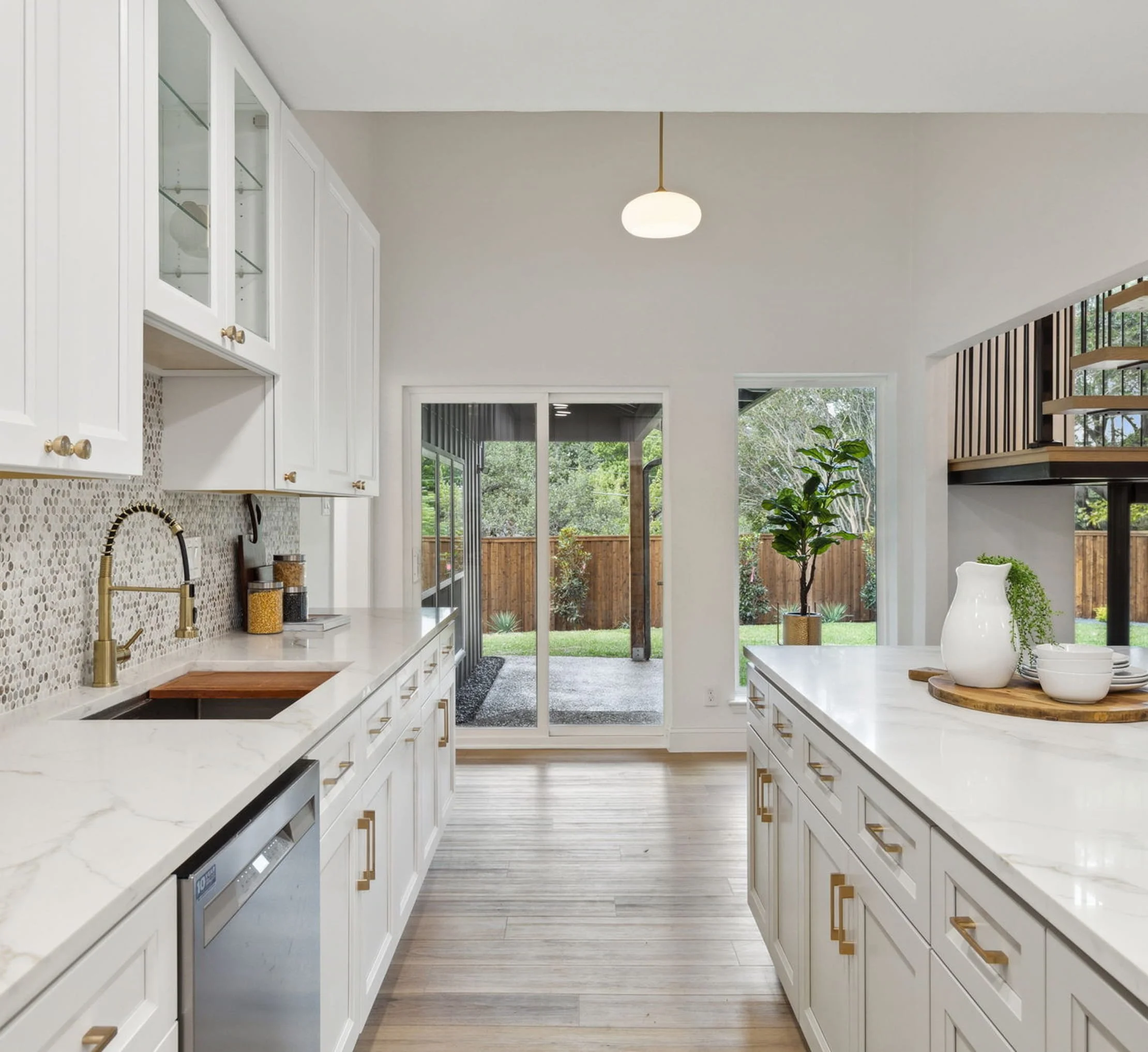 Modern kitchen with white cabinets, marble countertops, a mosaic tile backsplash, gold hardware, and large windows overlooking a backyard with trees.