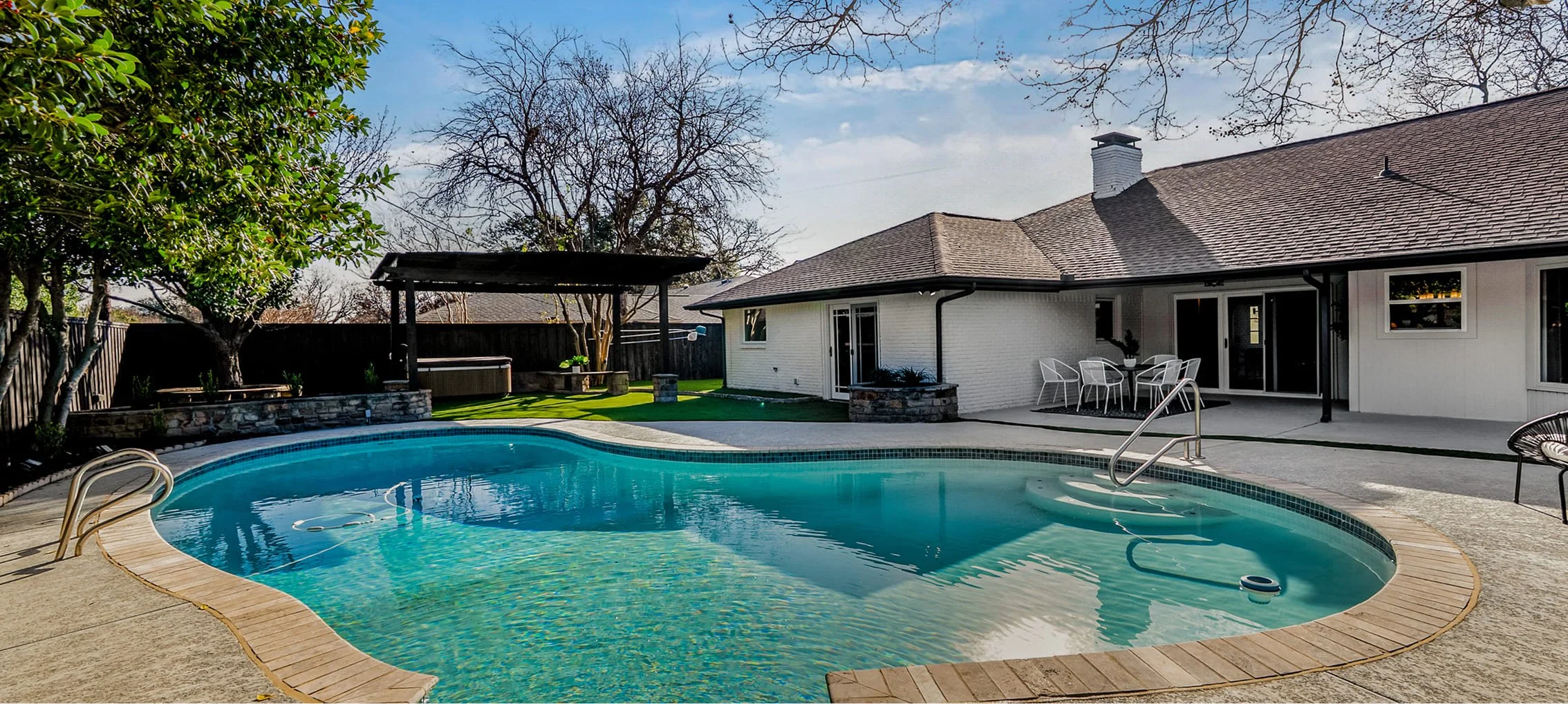 Backyard with a swimming pool, patio furniture, a covered hot tub, and a white house with a brown roof, surrounded by trees and a black fence, under a partly cloudy sky.