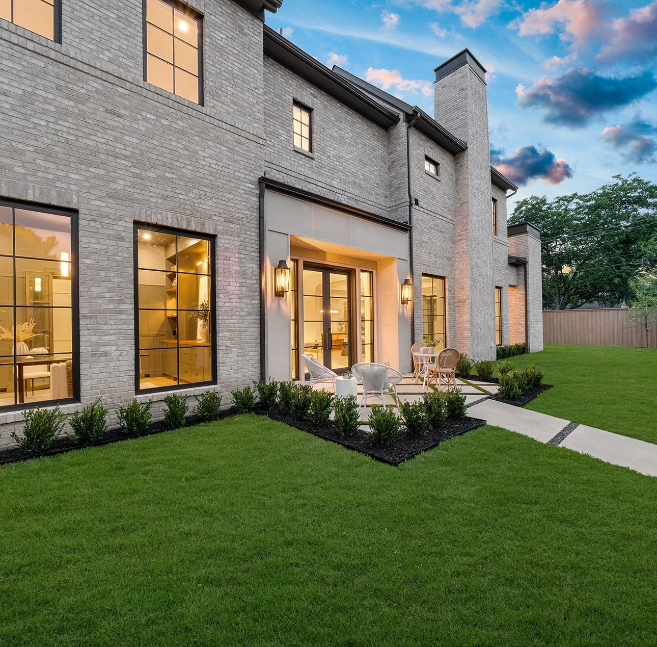 Modern brick house with large windows and a small backyard patio, illuminated by outdoor lights during dusk, with a well-maintained lawn and a wooden fence in the background.