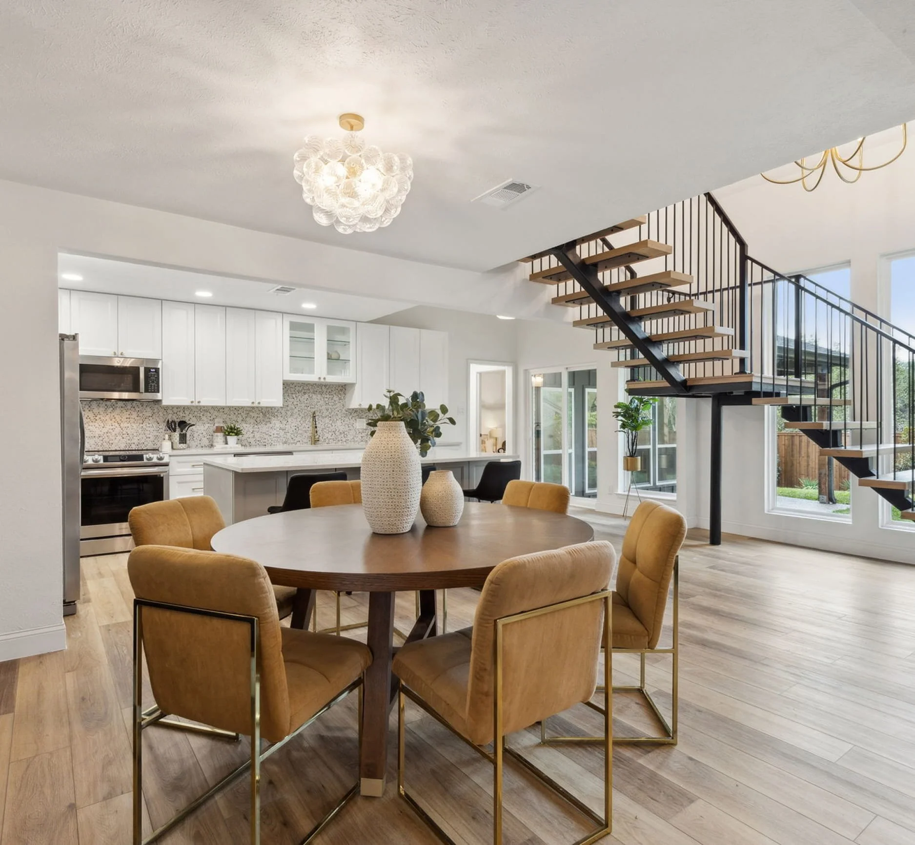 Open concept kitchen and dining area with hanging light fixture, staircase with wooden steps and black metal railing, and large windows.