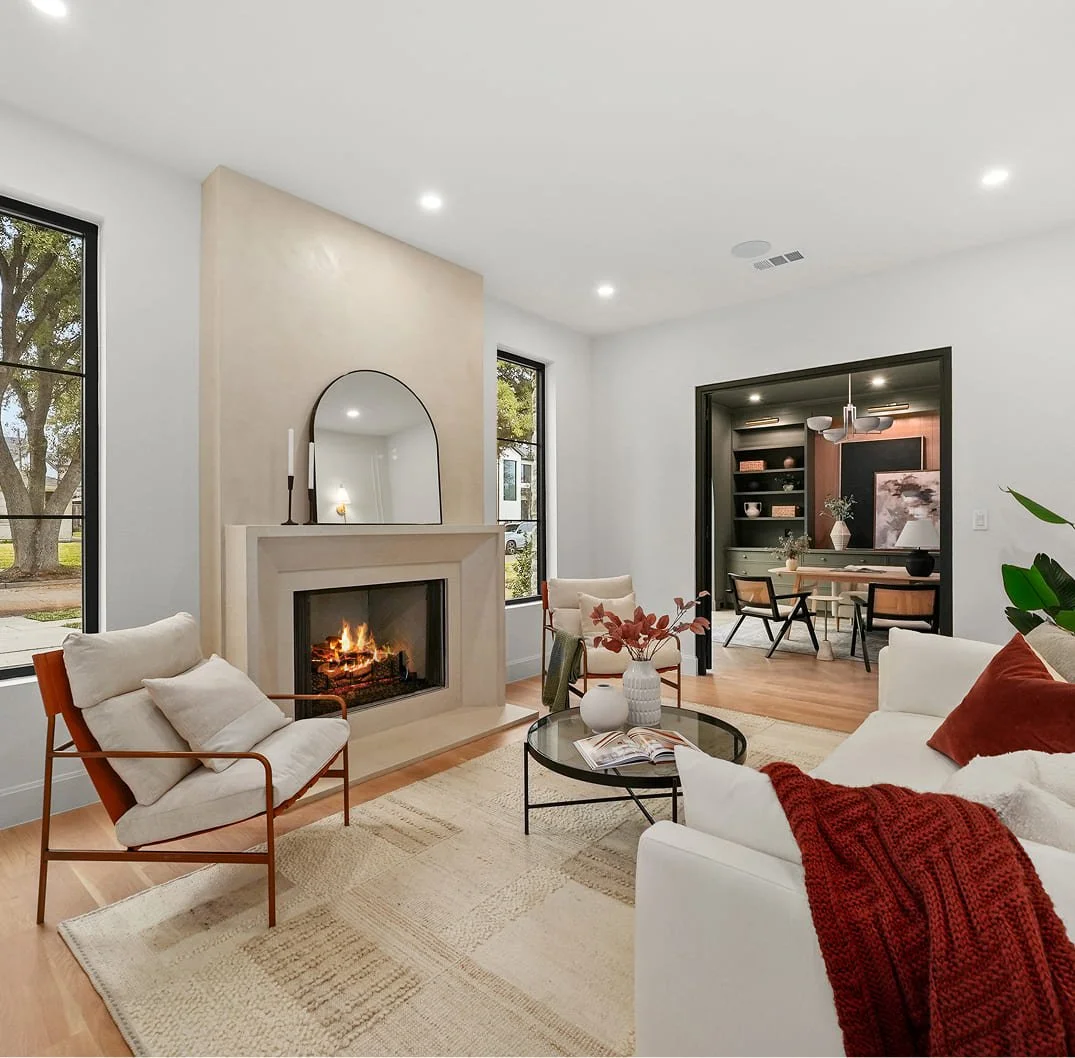 Modern living room with a fireplace, white sofa, beige armchair, glass coffee table, and large windows showing trees outside.