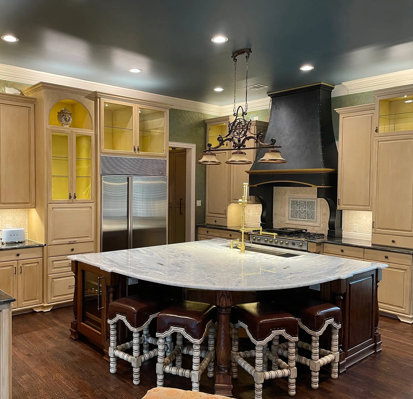 A spacious kitchen with cream-colored cabinetry, a large island with a white marble top, and a black range hood above a stove. A brass chandelier hangs over the island, and there are brown and white patterned stools around the table. The kitchen has warm lighting and wooden floors.