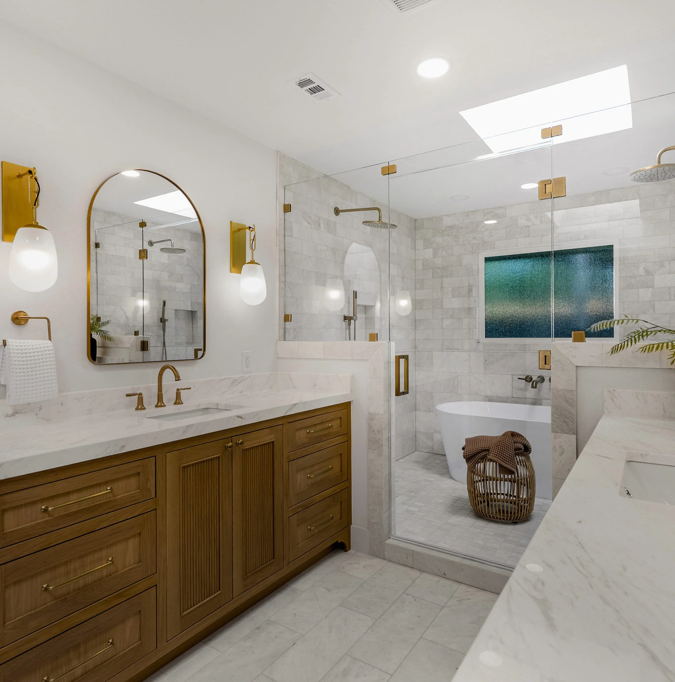 Modern bathroom with a large mirror, wall-mounted lights, a marble countertop sink, wooden vanity, walk-in shower with glass door, rain shower head, shower niche, window with frosted glass, and a wicker laundry basket.