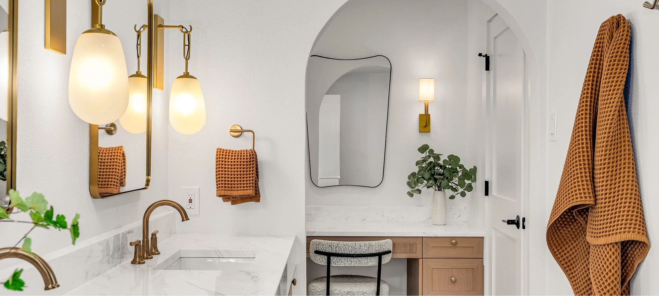 Modern bathroom with white walls, gold fixtures, a double sink with marble countertop, and orange towels hanging on wall hooks. There is a mirror, a wall-mounted light, and a small vanity area with a chair and a potted plant.