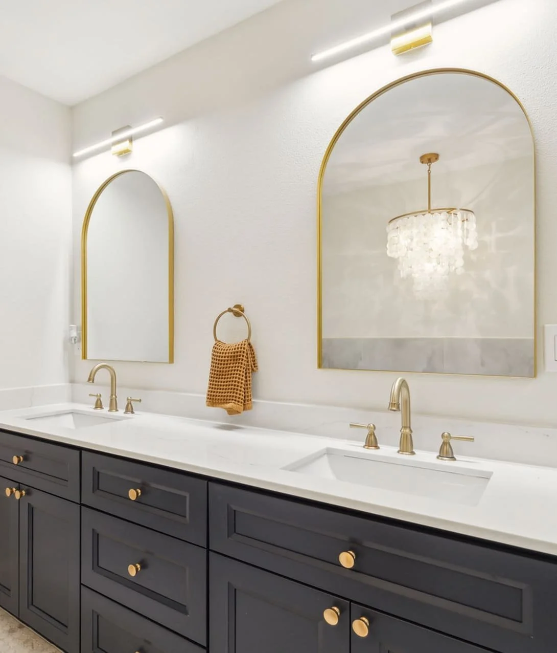 Modern bathroom with double vanity, white countertop, dark blue cabinets with gold knobs, two arched mirrors with gold frames, wall-mounted gold towel ring with brown towel, elegant chandelier reflected in one mirror, and minimal lighting.