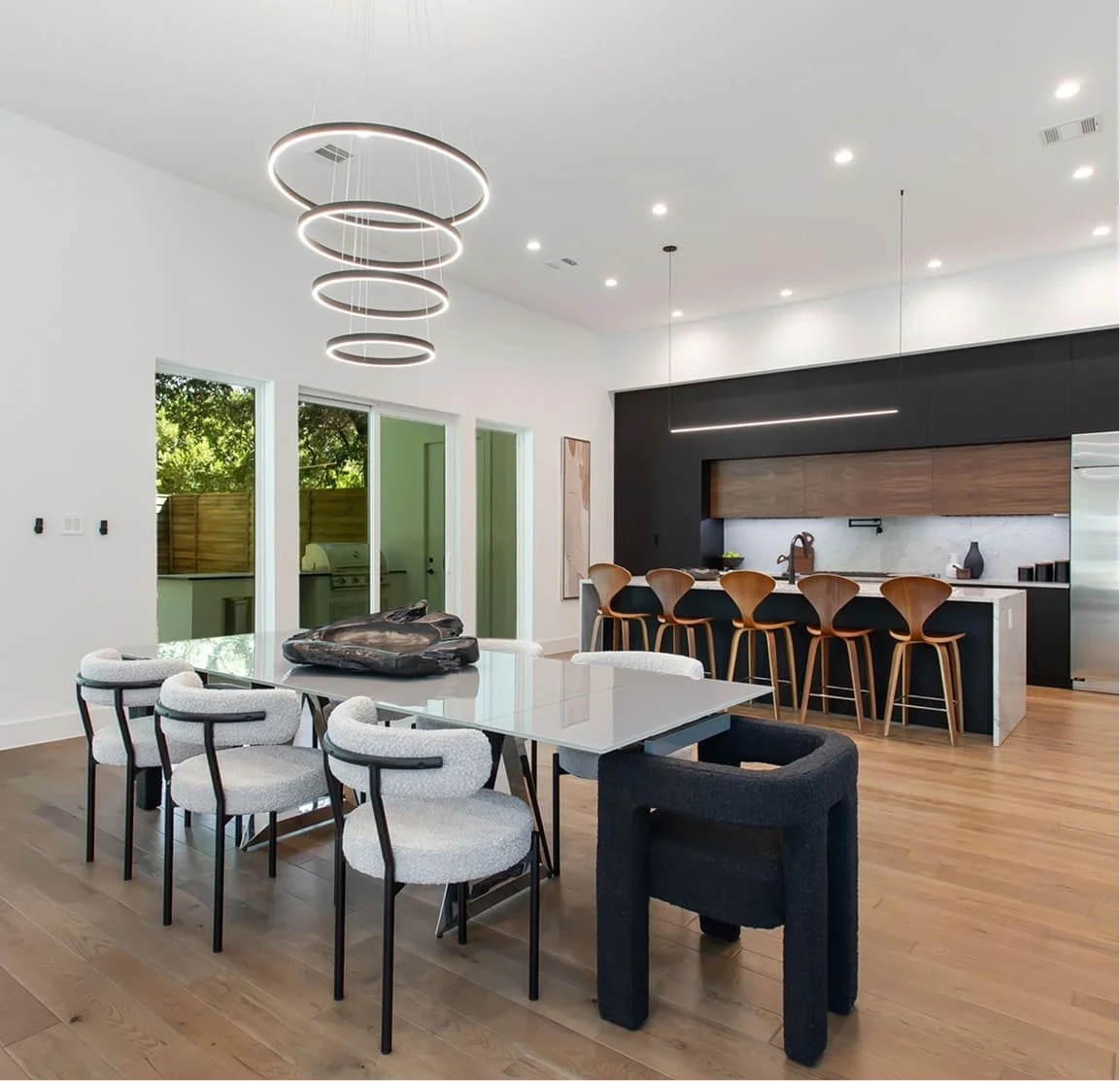 Modern open-concept kitchen and dining area with a white wall, wooden floors, and natural light. Features a white dining table with eight chairs, black and wood bar stools at a kitchen island, and contemporary ceiling lighting.