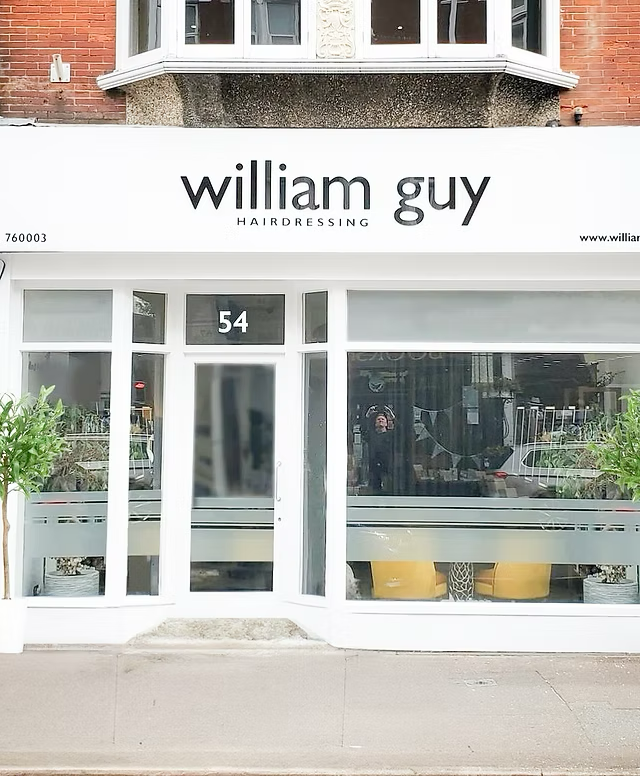 storefront sign for William Guy Hairdressing at 54 with large front window, two small trees flanking the entrance, and a reflection of the street in the glass.