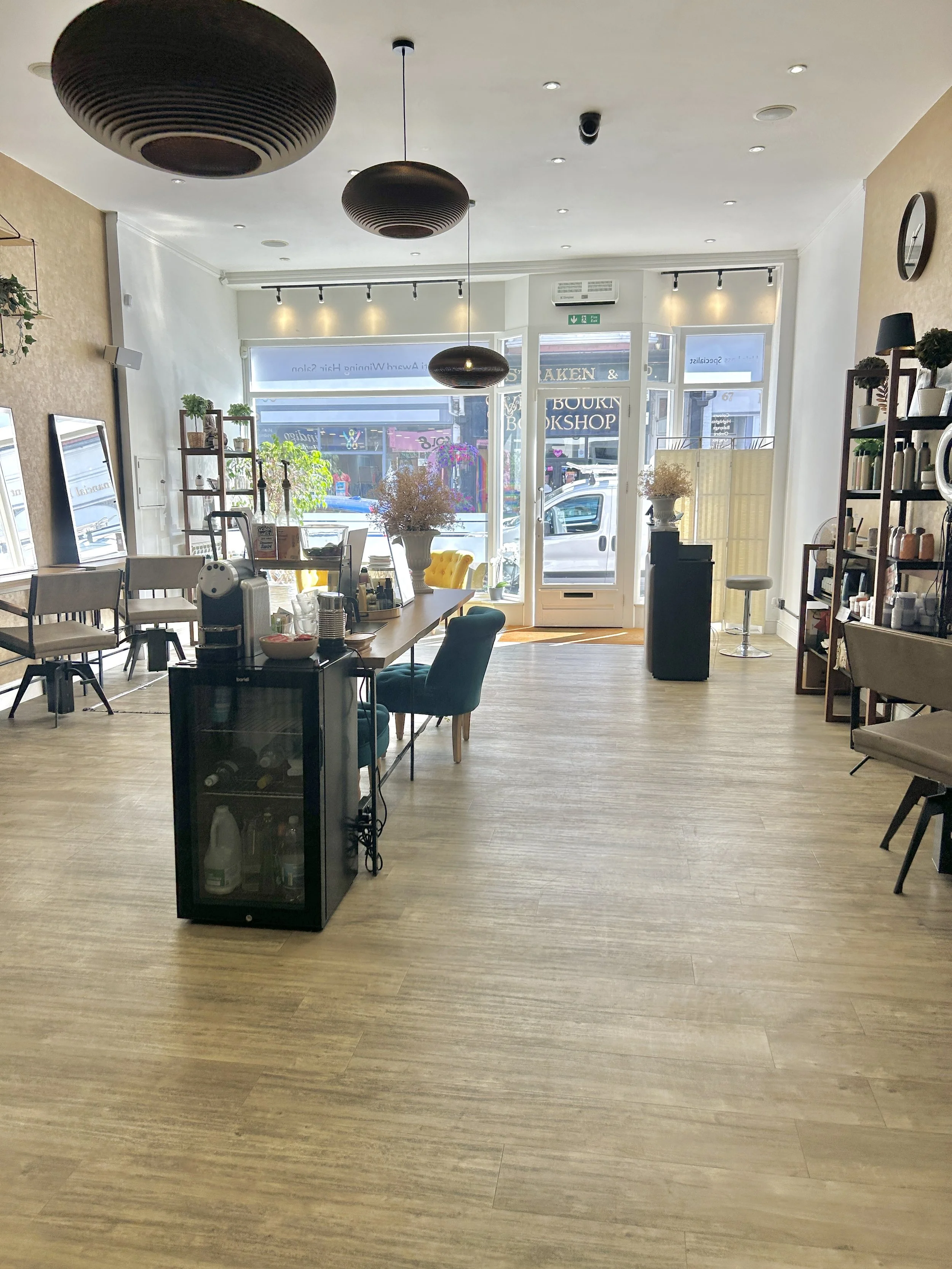 Bright and airy interior of a cafe or shop with large front windows, wooden flooring, a black refrigerator with bottles, chairs, shelves with plants and products, and decorative lighting fixtures.