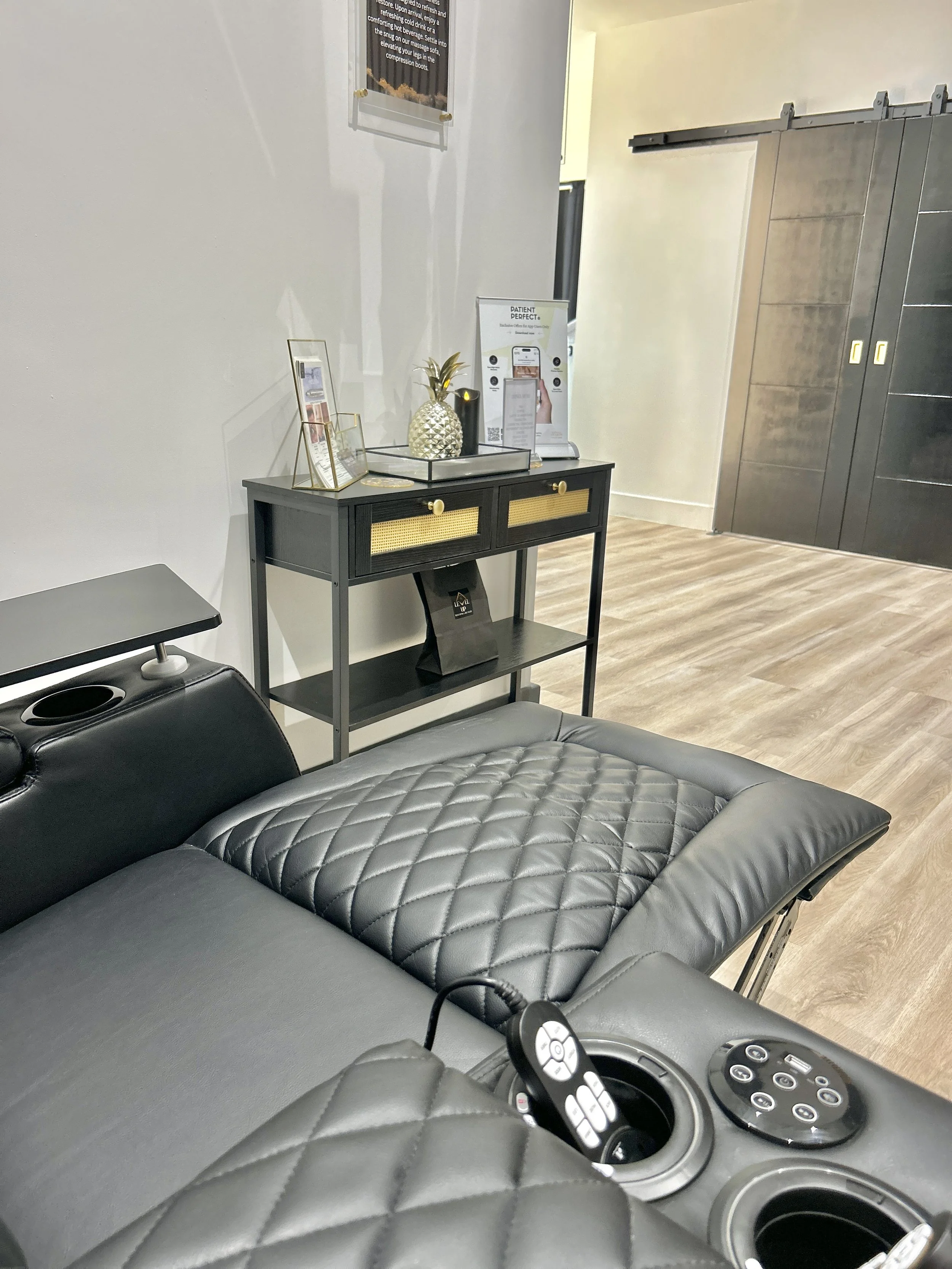 Interior of a medical or spa facility with a leather examination or massage table, a small black cart with decorative items including a pineapple-shaped ornament, and a door with a sliding mechanism. There are informational posters on the wall.