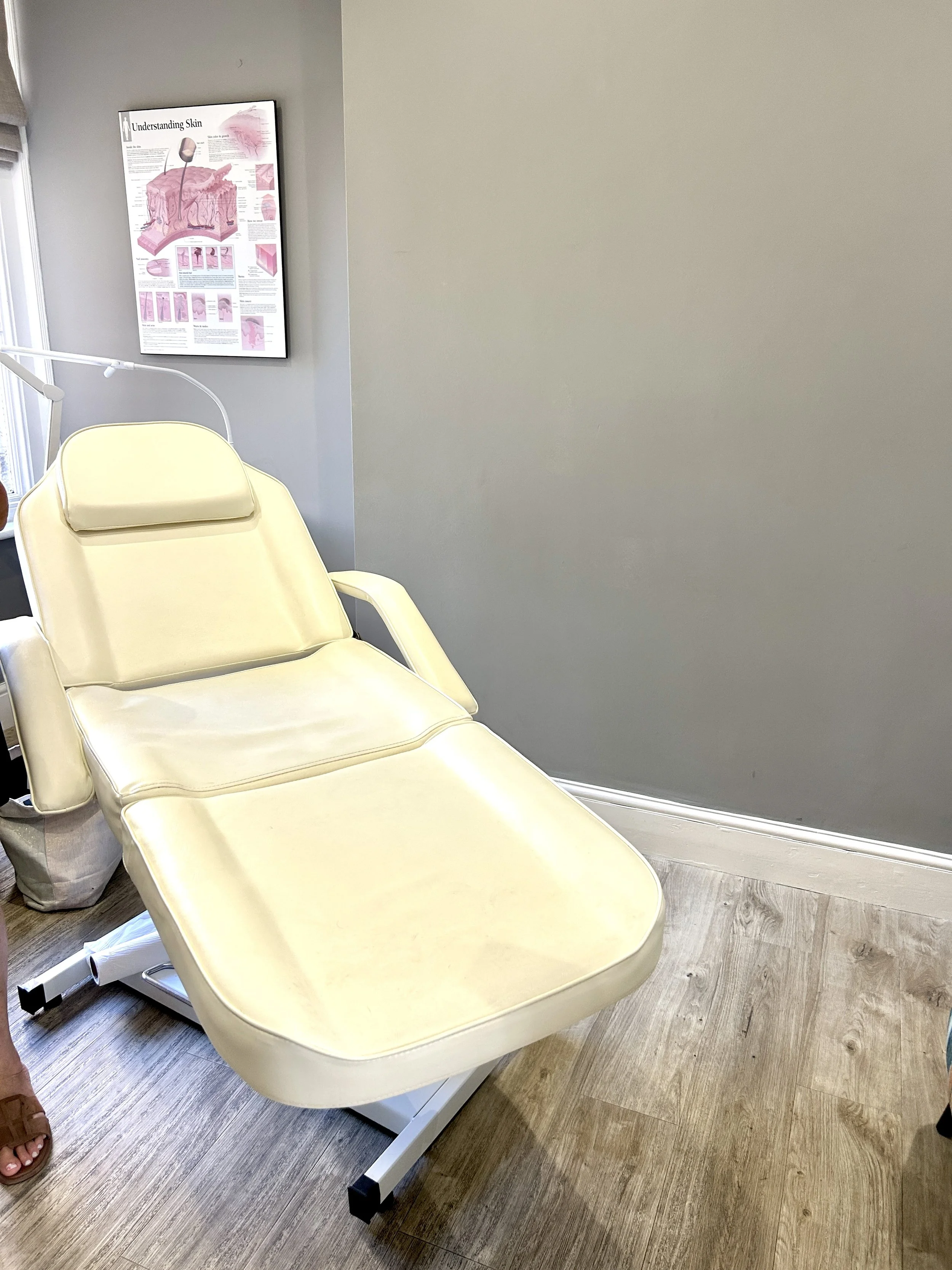 Medical examination room with a beige adjustable examination table and a chart on the wall showing skin layers.