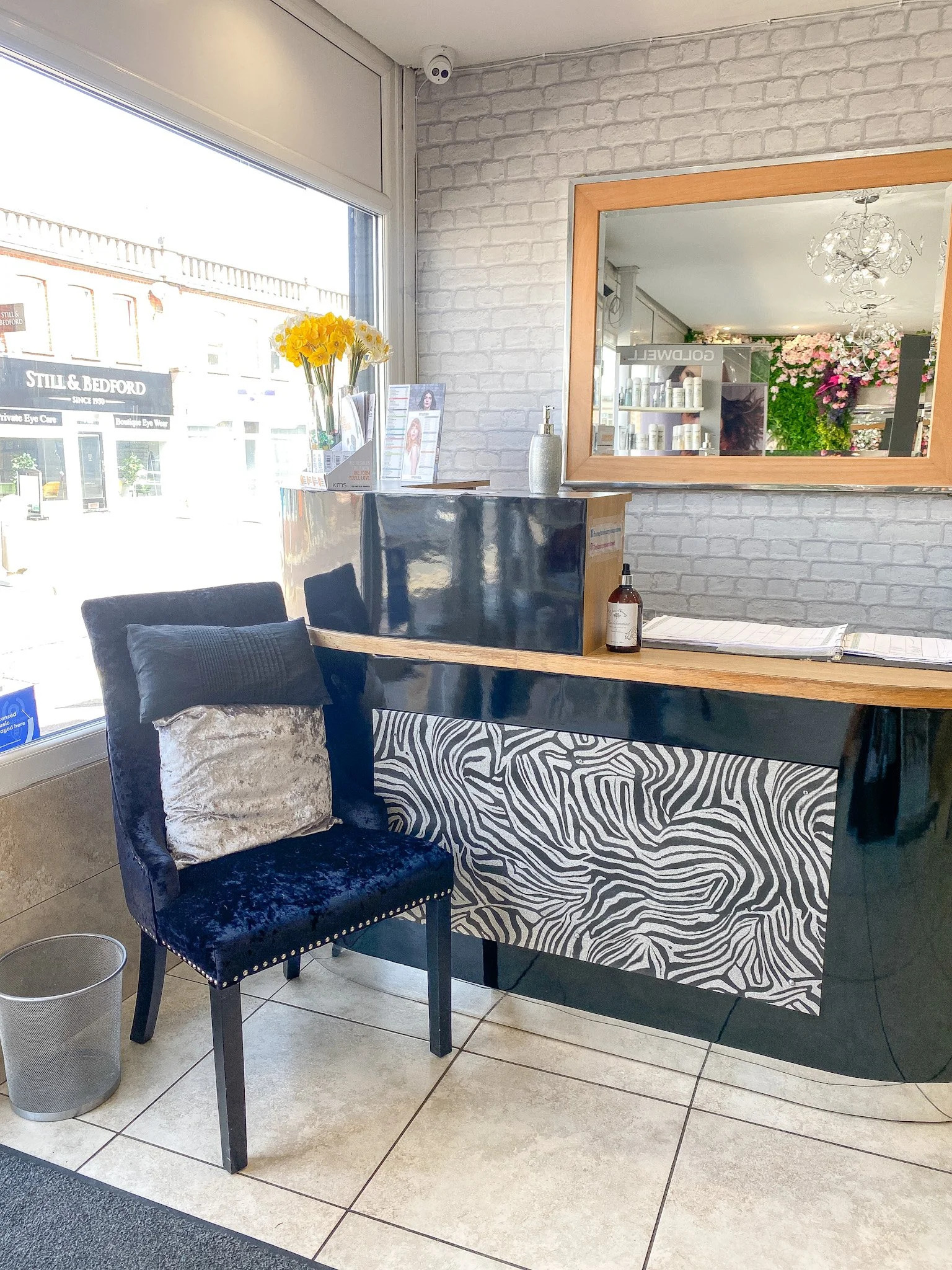Inside a modern hair salon with a white brick wall, a mirror, a black velvet chair with a beige pillow, a small wooden table with a zebra print cloth, a soap dispenser, hair products, and a window with a view of storefronts outside.