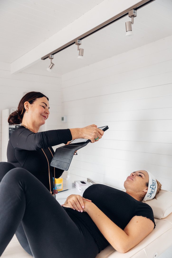 A woman lying on a treatment bed with her hands on her stomach, wearing a headband labeled 'SkinPen,' faces a smiling woman standing nearby. The standing woman is holding a device and a clipboard, in a clinical setting with bright white walls and ceiling.