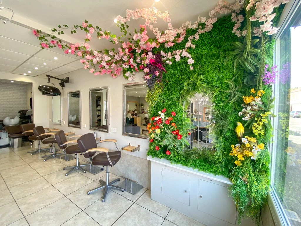 Hair salon interior with styling chairs along the wall, mirrors, and a large window decorated with a vibrant green wall filled with pink, purple, white, and yellow flowers and greenery.