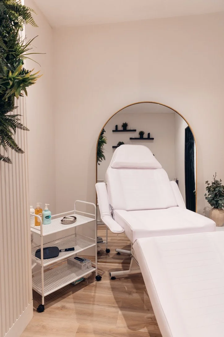 A medical treatment room with a white adjustable examination chair, a large mirror behind the chair, a white rolling cart with medical supplies, and potted plants on shelves and the floor.