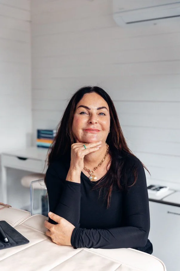 A woman with long dark hair, wearing a black top and jewelry, sitting at a desk in a bright office, smiling and resting her chin on her hand.