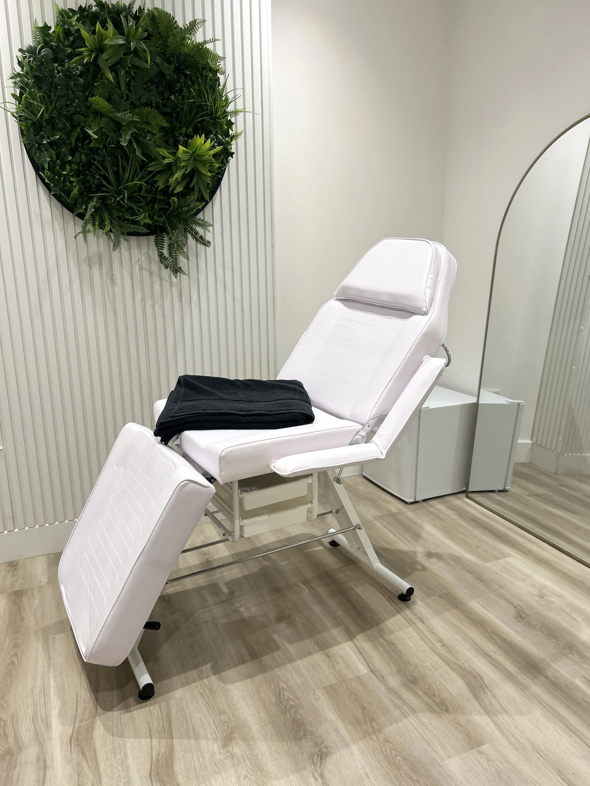 A white medical chair with a headrest, covered with a black cloth, set in a skincare room with light wood flooring and a textured white wall.